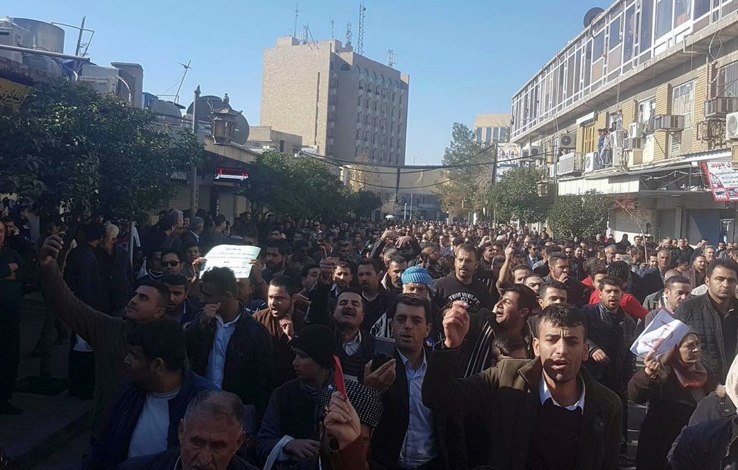 The image depicts a large crowd of people gathered on a street, likely participating in a protest or demonstration. The individuals appear to be expressing their views passionately, with some holding signs. The setting includes buildings and urban infrastructure, indicating the event is taking place in a city. The atmosphere seems intense, with many people visible in the foreground and background, suggesting a significant turnout for the event.