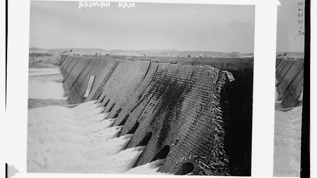 L'image représente un barrage, probablement le barrage d'Assouan, avec une structure en béton. On peut voir de l'eau s'écoulant à travers des ouvertures sur le côté du barrage. Le paysage environnant semble naturel, et le ciel est clair. L'image évoque une impression de puissance hydraulique et d'ingénierie.