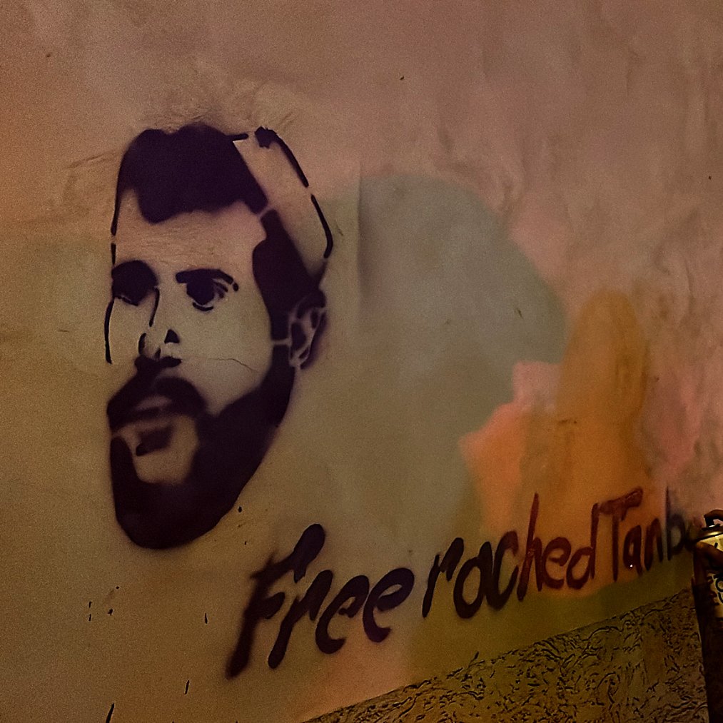 L'image montre une personne de dos, habillée d'un sweat à capuche blanc, en train de peindre un graffiti sur un mur. Le graffiti représente le visage d'un homme, probablement une figure connue, avec la phrase "Free roched Tank" écrite en dessous. La scène se déroule la nuit, et l'éclairage crée des reflets colorés sur le mur. On peut également apercevoir un portail ou une autre surface à proximité avec un autre graffiti en arrière-plan.