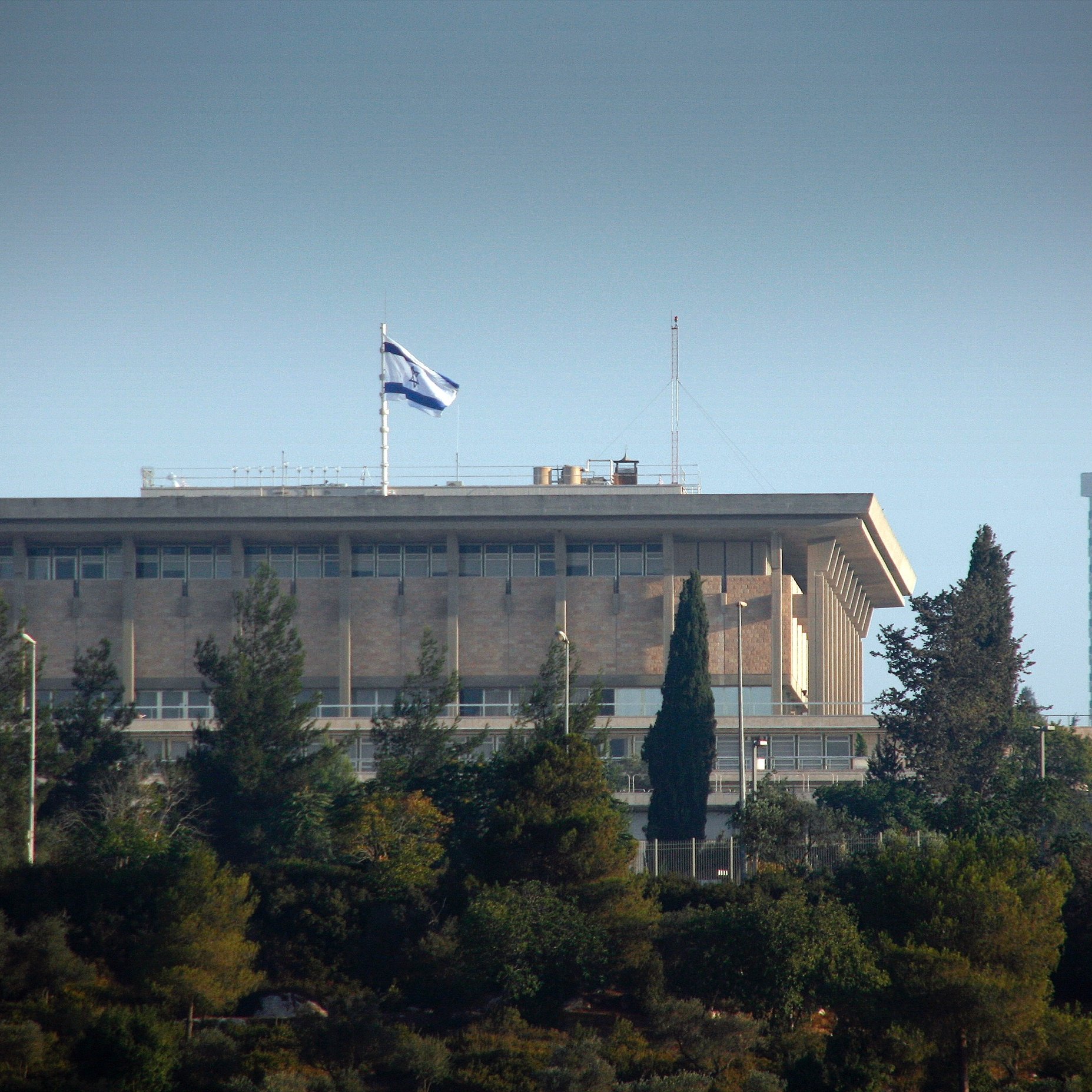 L'image montre un bâtiment imposant, probablement un bâtiment gouvernemental, situé sur une colline. On peut voir le drapeau d'Israël flotter au sommet du bâtiment. Celui-ci est entouré de verdure et d'arbres, indiquant que l'environnement est naturel et paisible. La structure a un style architectural moderne avec de grandes fenêtres et des balcons. L'éclairage suggère que la photo a été prise pendant la journée.