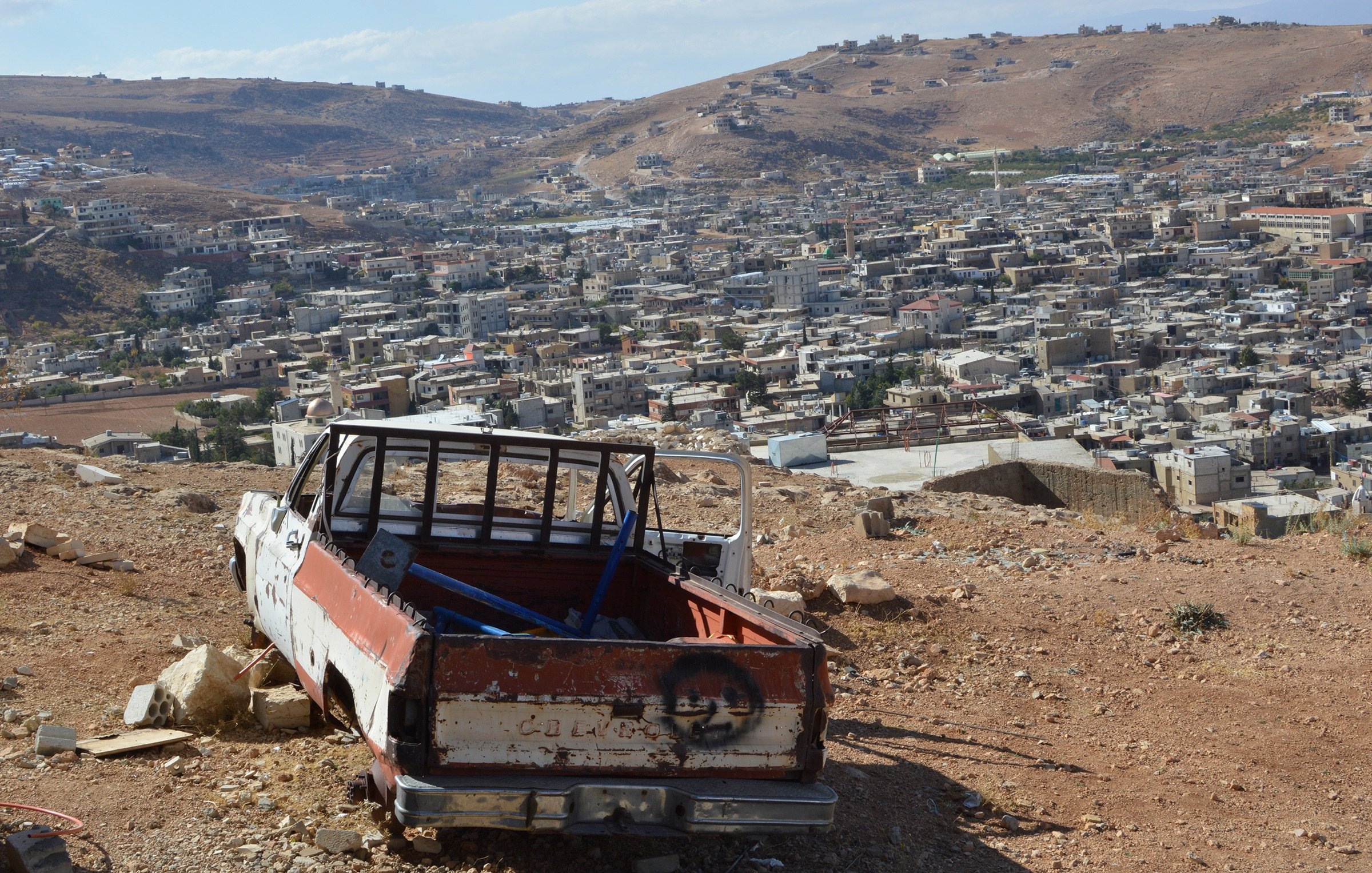 L'image montre une scène de paysage où un ancien véhicule, probablement une camionnette, est abandonné sur une colline. La camionnette est en mauvais état, avec une partie de la carrosserie rouillée et peinte en blanc et rouge. En arrière-plan, on peut voir un village ou une ville avec de nombreuses maisons, s'étendant sur les collines. Le ciel est partiellement nuageux, et le paysage environnant est aride, typique des terrains montagneux.