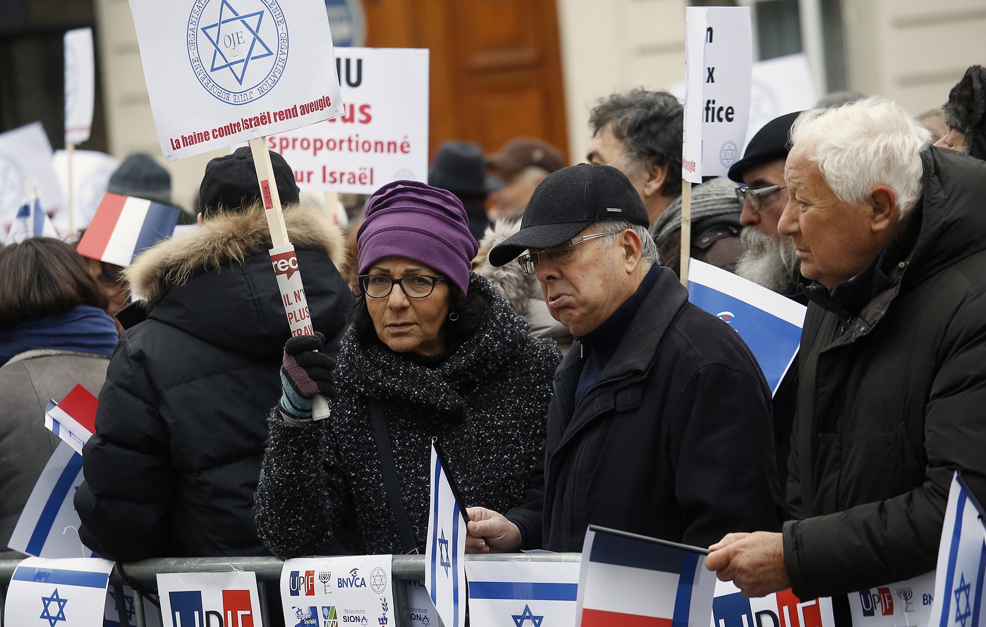 L'image montre une manifestation où un groupe de personnes tient des pancartes et des drapeaux. Les participants semblent se rassembler pour exprimer leurs opinions sur un sujet lié à Israël. Certains portent des signes indiquant des messages clairs, tandis que d'autres affichent des drapeaux français. L'atmosphère semble sérieuse et déterminée.