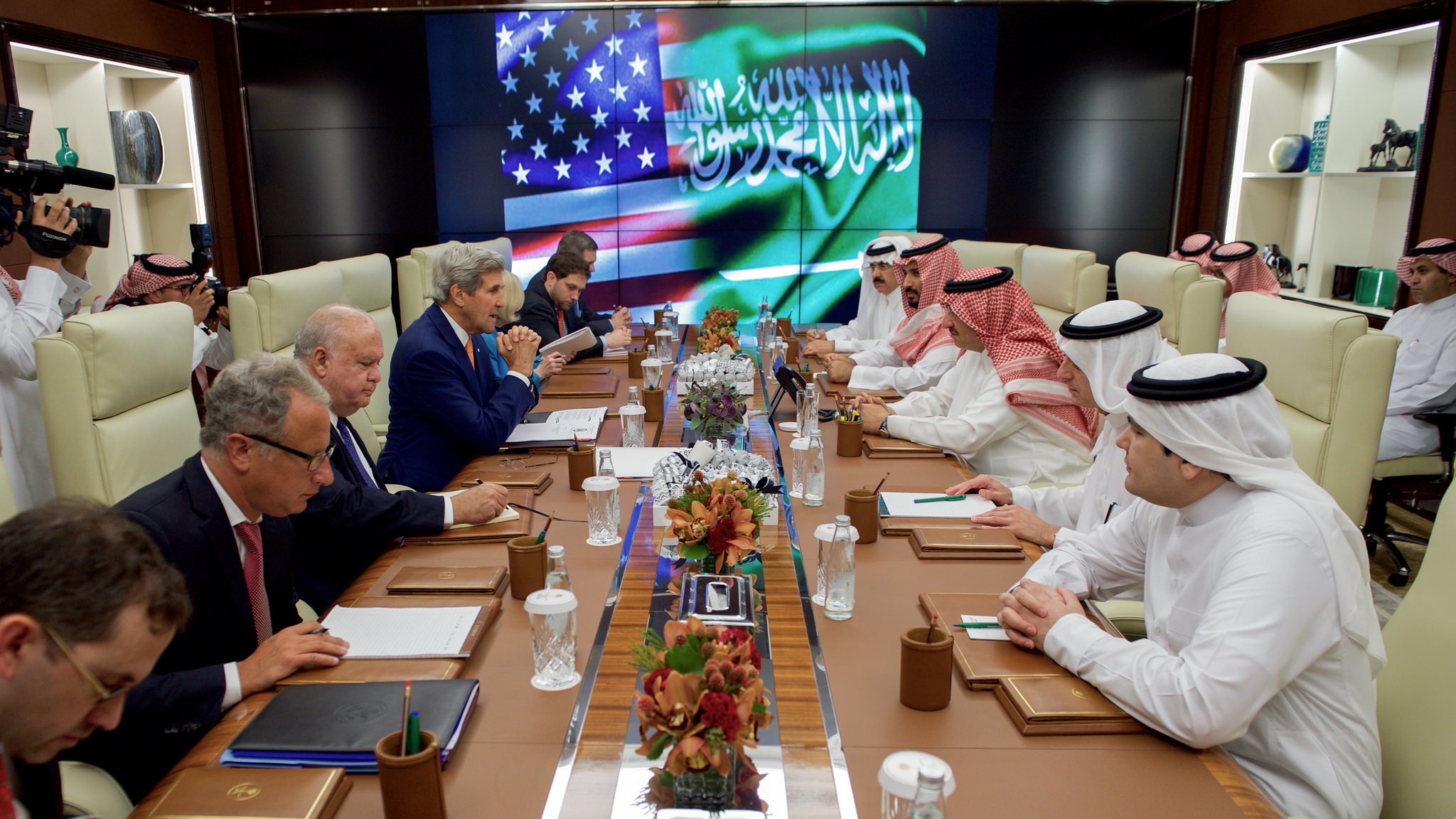 تظهر الصورة اجتماعًا رسميًا يجمع بين مسؤولين سعوديين وأمريكيين. في غرفة تجمعهم، يتواجد ممثلون من كلا الجانبين، حيث يجلس الرجلان في المقدمة على طاولة مستديرة، بينما يقف آخرون بجانبهم. هناك شاشة في الخلف تعرض علمي السعودية والولايات المتحدة، مما يعكس شراكة أو تعاون بين البلدين. يبدو أن الجميع يركز على المناقشات الجارية مع وجود ملاحظات وأدوات على الطاولة.