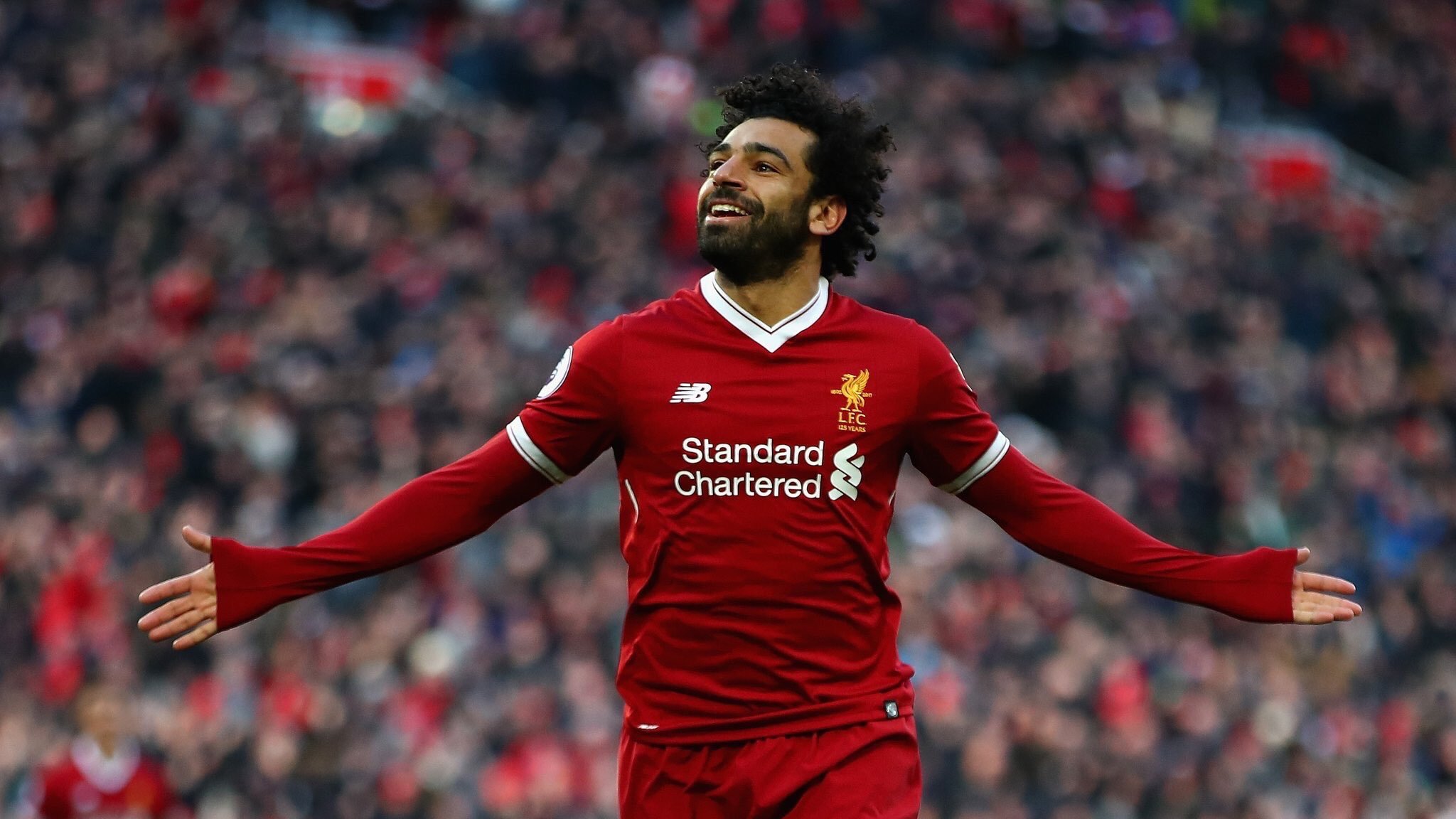 L'image montre un footballeur célébrant un but. Il porte un maillot rouge avec le logo de Liverpool FC, ainsi que des logos de sponsors. Son expression est joyeuse, avec un grand sourire, et il étend les bras, manifestant sa joie devant les supporters. L'arrière-plan est flou, mais on peut deviner une foule enthousiaste dans un stade.