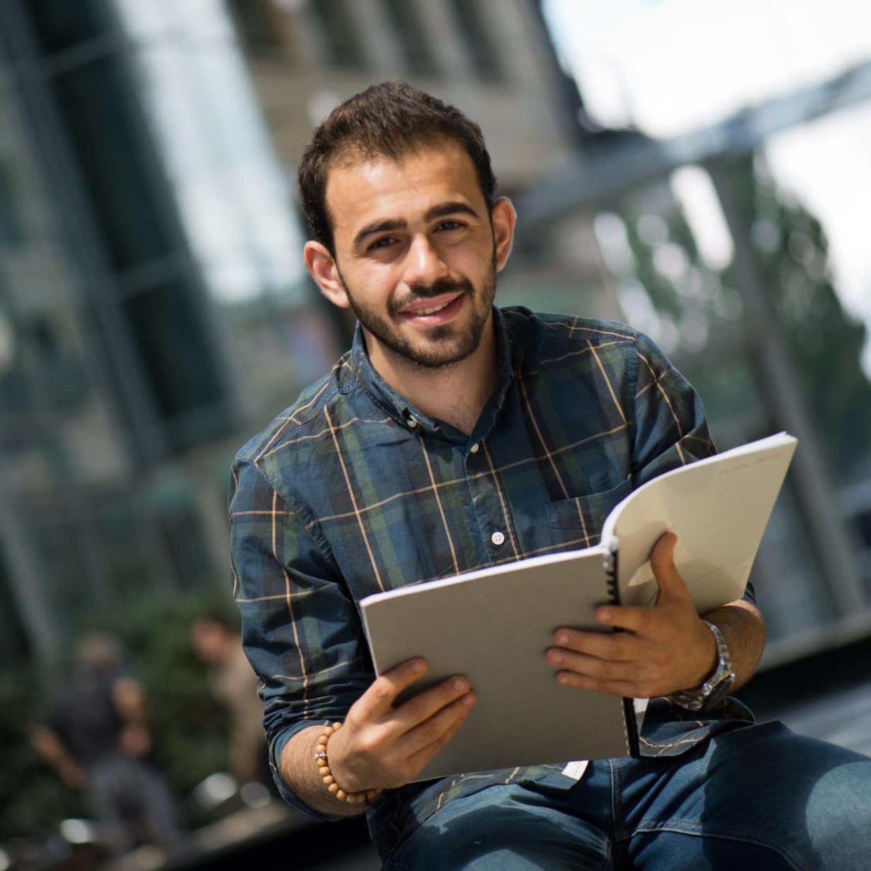 The image features a young man sitting outdoors, holding an open notebook or binder. He has a friendly smile and is wearing a plaid shirt. The background includes modern architecture and greenery, suggesting an urban setting. The atmosphere appears bright and inviting, indicating a pleasant day.