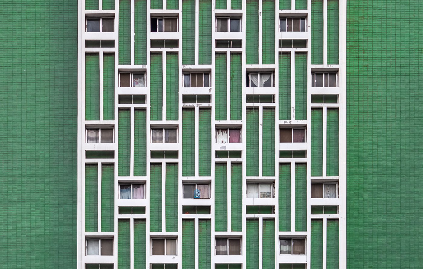 L'image montre la façade d'un immeuble aux fenêtres disposées de manière régulière. Les murs sont peints en vert, créant un contraste avec les cadres des fenêtres, qui sont de couleur blanche. Les fenêtres sont variées en termes d'ouverture, certaines étant complètement ouvertes et d'autres fermées, laissant entrevoir quelques éléments de la vie intérieure, comme des rideaux ou des objets. L'ensemble présente un aspect géométrique et moderne.