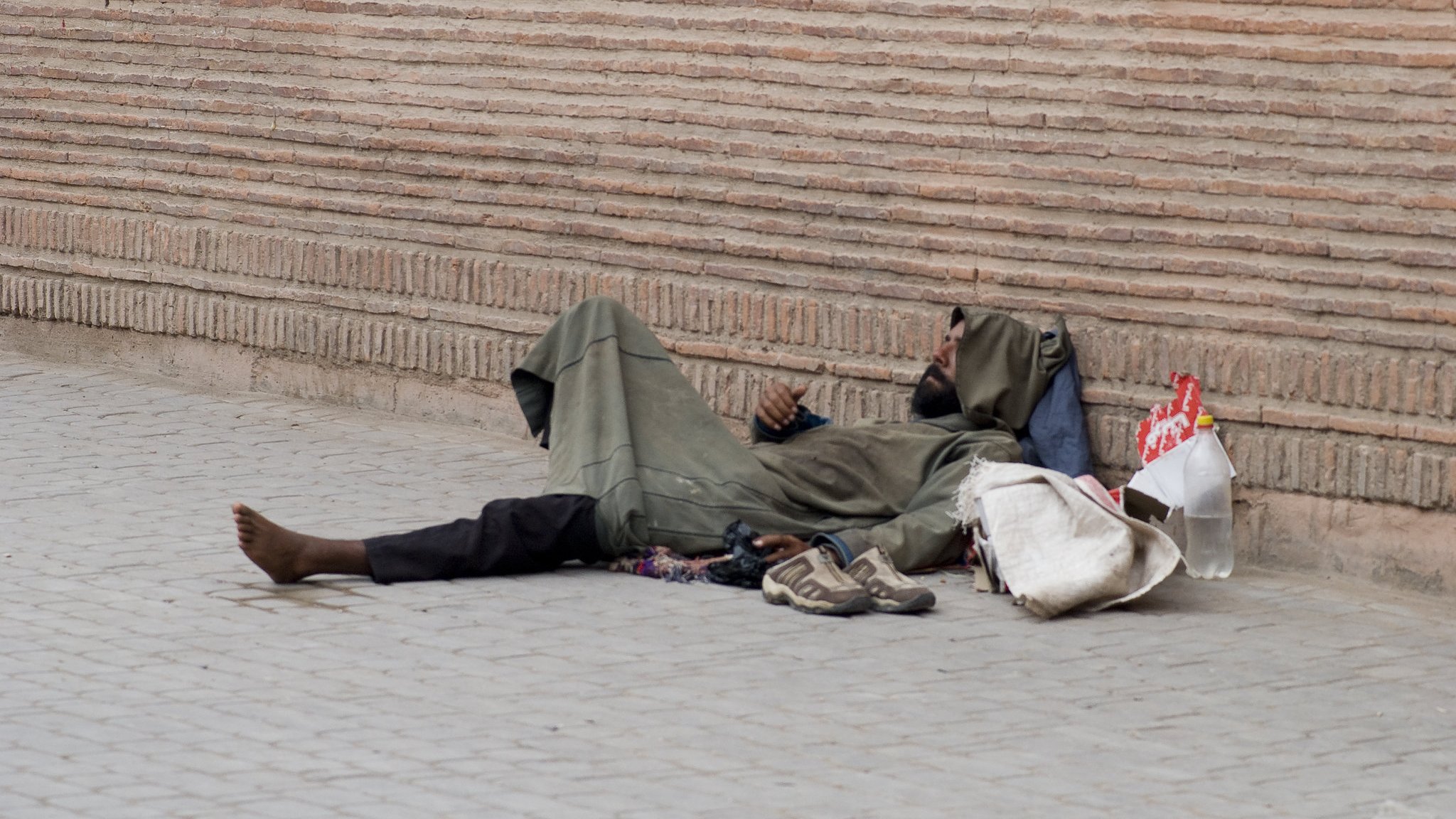 L'image montre une personne allongée sur le sol, semblant sans abri. Elle porte des vêtements sombres et usés, et se trouve à côté d'une paroi de briques. On voit des objets éparpillés autour d'elle, comme des chaussures et un sac. L'environnement semble urbain et le ton de l'image est contemplatif.