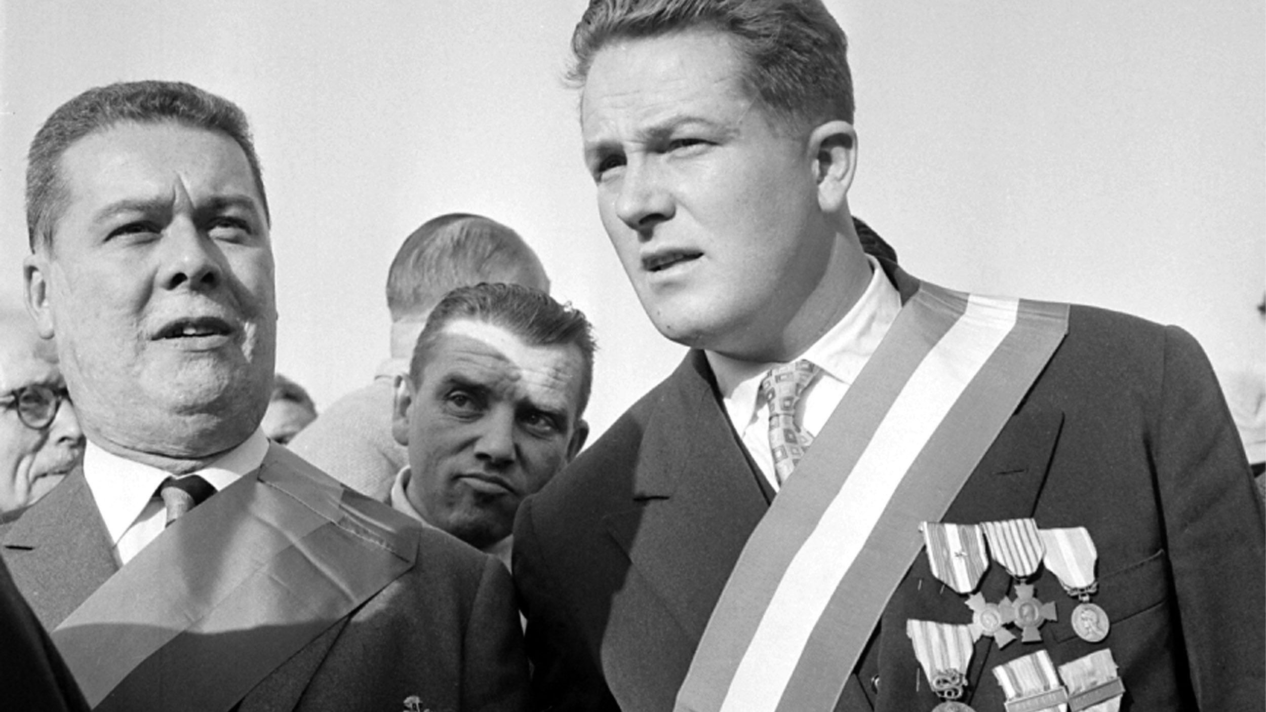 L'image montre deux hommes qui semblent participer à un événement officiel ou une cérémonie. L'un des hommes porte plusieurs médailles accrochées à sa veste, ainsi qu'une écharpe. L'autre homme porte également une écharpe, mais il a moins de médailles visibles. L'ambiance paraît solennelle, avec d'autres personnes en arrière-plan. Les expressions des deux hommes suggèrent qu'ils sont engagés dans une conversation ou qu'ils se concentrent sur l'événement.