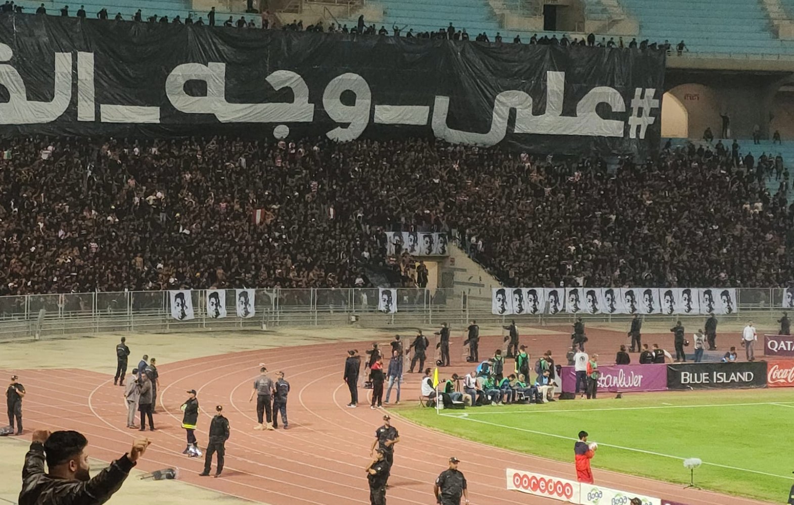 A large crowd at a stadium with a black banner and fans cheering passionately.