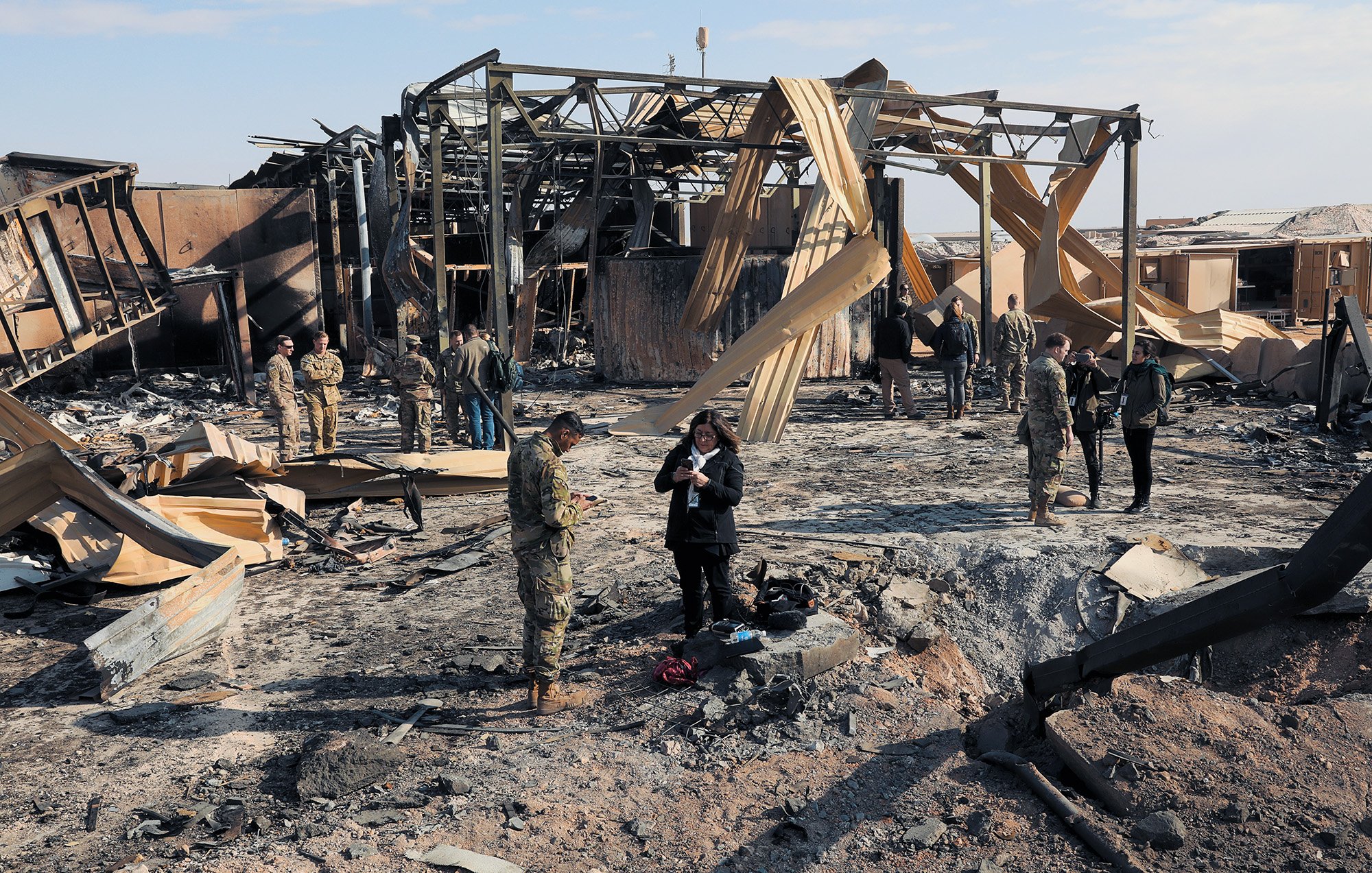 در این تصویر، صحنه‌ای از یک منطقه آسیب‌دیده به‌دلیل آتش‌سوزی یا انفجار دیده می‌شود. ساختمان‌ها به شدت خراب شده‌اند و بخشی از سازه‌ها و دیواره‌ها به شکل ناپایداری در حال افتادن هستند. چند نفر از نیروهای نظامی و خبرنگاران در میان خرابه‌ها در حال بررسی وضعیت و جمع‌آوری اطلاعات هستند. زمین پوشیده از خاک و بقایای سوخته است و آسمان آبی و صاف به نظر می‌رسد.