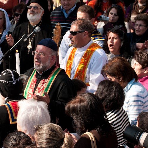 L'image représente un groupe de personnes rassemblées lors d'une cérémonie religieuse. Au centre, des figures cléricales en vêtements liturgiques colorés chantent et prient, entourées de fidèles. On aperçoit des chants, des bougies et des fleurs décoratives, ce qui suggère un événement festif ou sacré. L'ambiance est chaleureuse et communautaire, avec une multitude de visages exprimant la dévotion.