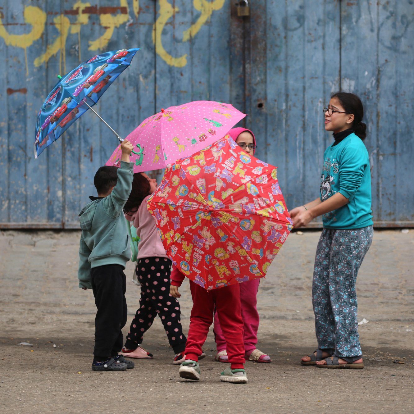 L'image montre un groupe d'enfants jouant dans la rue. Ils ont des parapluies colorés, certains avec des motifs amusants. Les enfants semblent joyeux et engagés dans leur jeu. En arrière-plan, il y a un mur bleu, qui donne une ambiance urbaine à la scène. Ils portent des vêtements confortables, adaptés à une activité en extérieur. L'atmosphère générale est légère et pleine de vie.