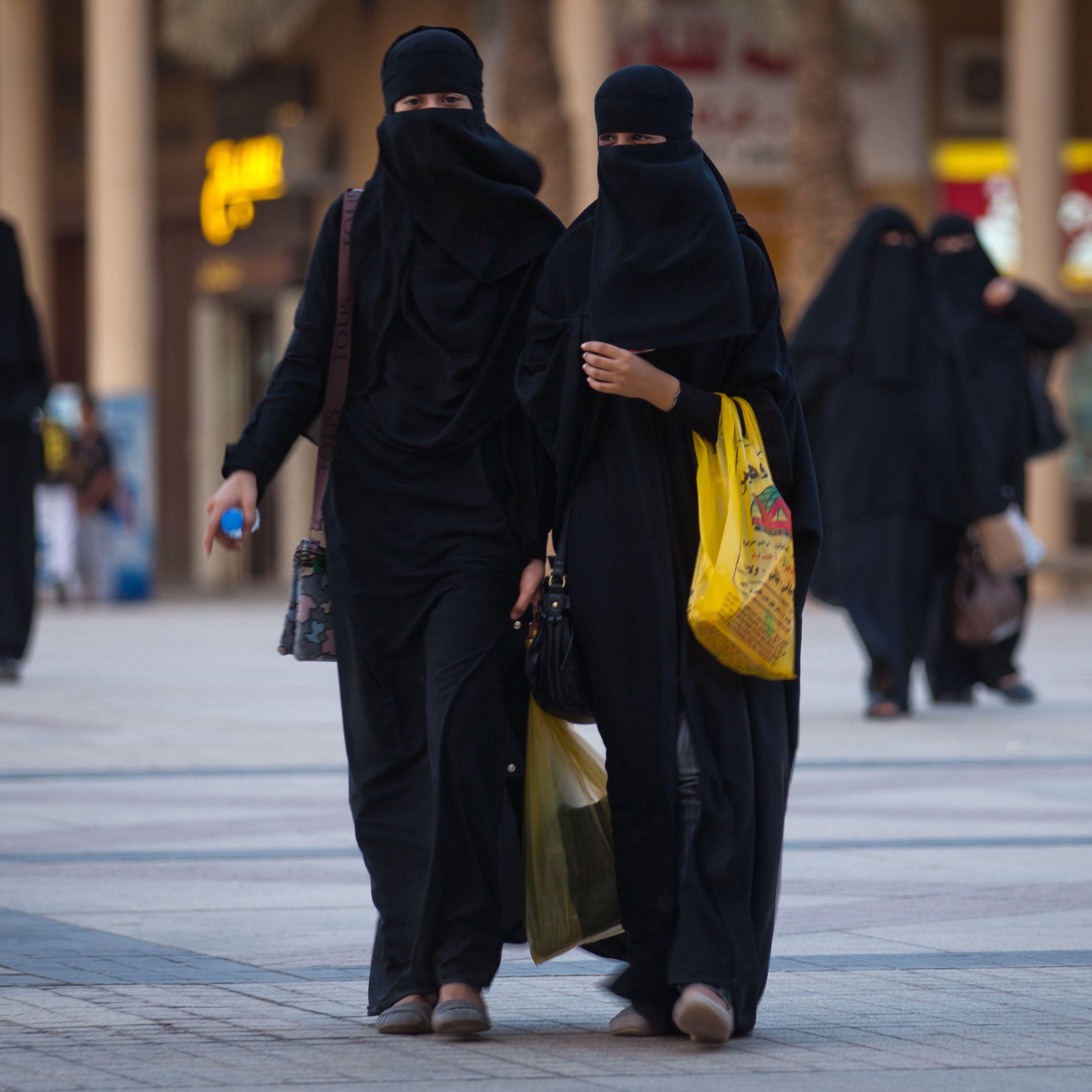 L'image montre deux femmes marchant côte à côte dans une zone urbaine. Elles portent des vêtements noirs, y compris des niqabs qui couvrent leur visage, laissant seulement les yeux visibles. L'environnement semble animé, avec des bâtiments en arrière-plan et d'autres personnes se déplaçant dans la même direction. L'une des femmes tient un sac, tandis que l'autre semble tenir un objet dans sa main. L'éclairage et l'architecture suggèrent un cadre contemporain.