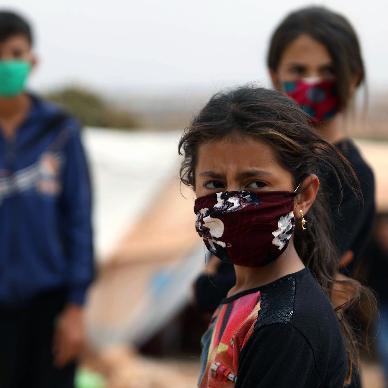 L'image montre un groupe d'enfants, dont une fille au premier plan, portant des masques. La jeune fille a les cheveux bruns et des yeux expressifs, semblant préoccupée. En arrière-plan, d'autres enfants se tiennent, également masqués, dans un environnement qui semble être un camp. L'atmosphère semble chargée d'émotion et de défi. Les couleurs des masques varient, ajoutant une touche de diversité visuelle à la scène.