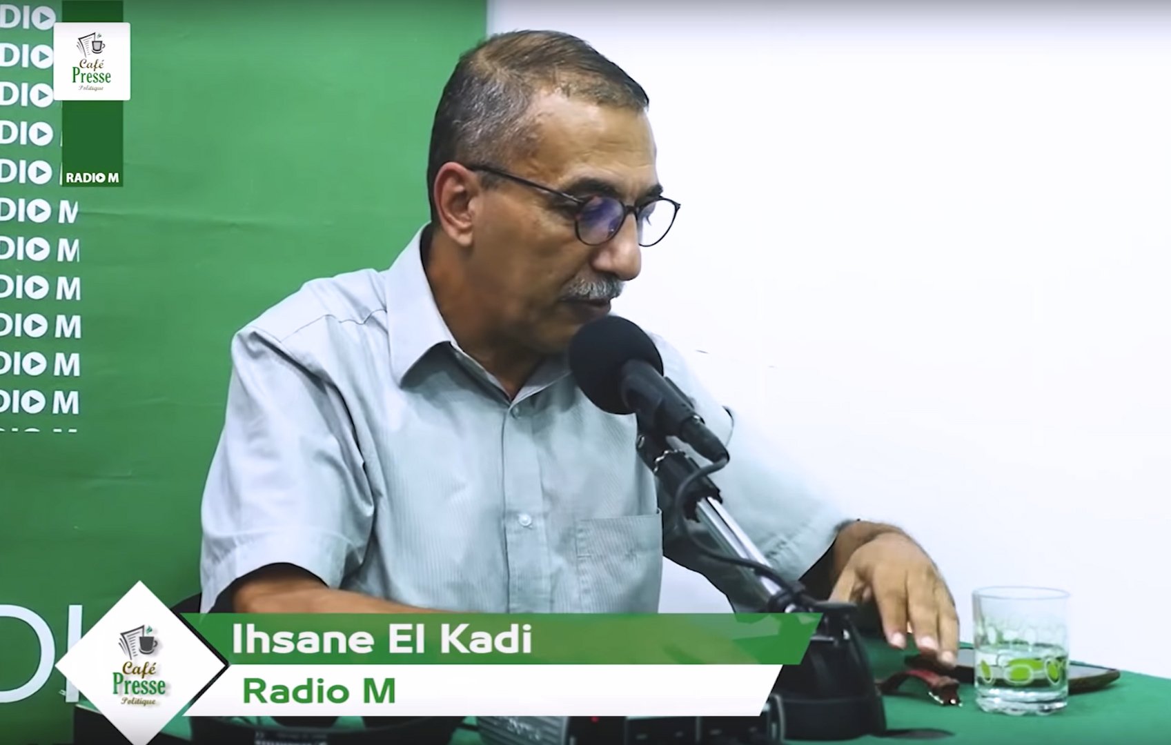 L'image montre un homme assis derrière un micro dans un studio de radio. Il porte une chemise à manches courtes et des lunettes. Sur la table devant lui, il y a un verre d'eau et un téléphone. L'arrière-plan est vert avec des éléments de branding pour "Radio M". Le nom "Ihsane El Kadi" est affiché en bas de l'image.