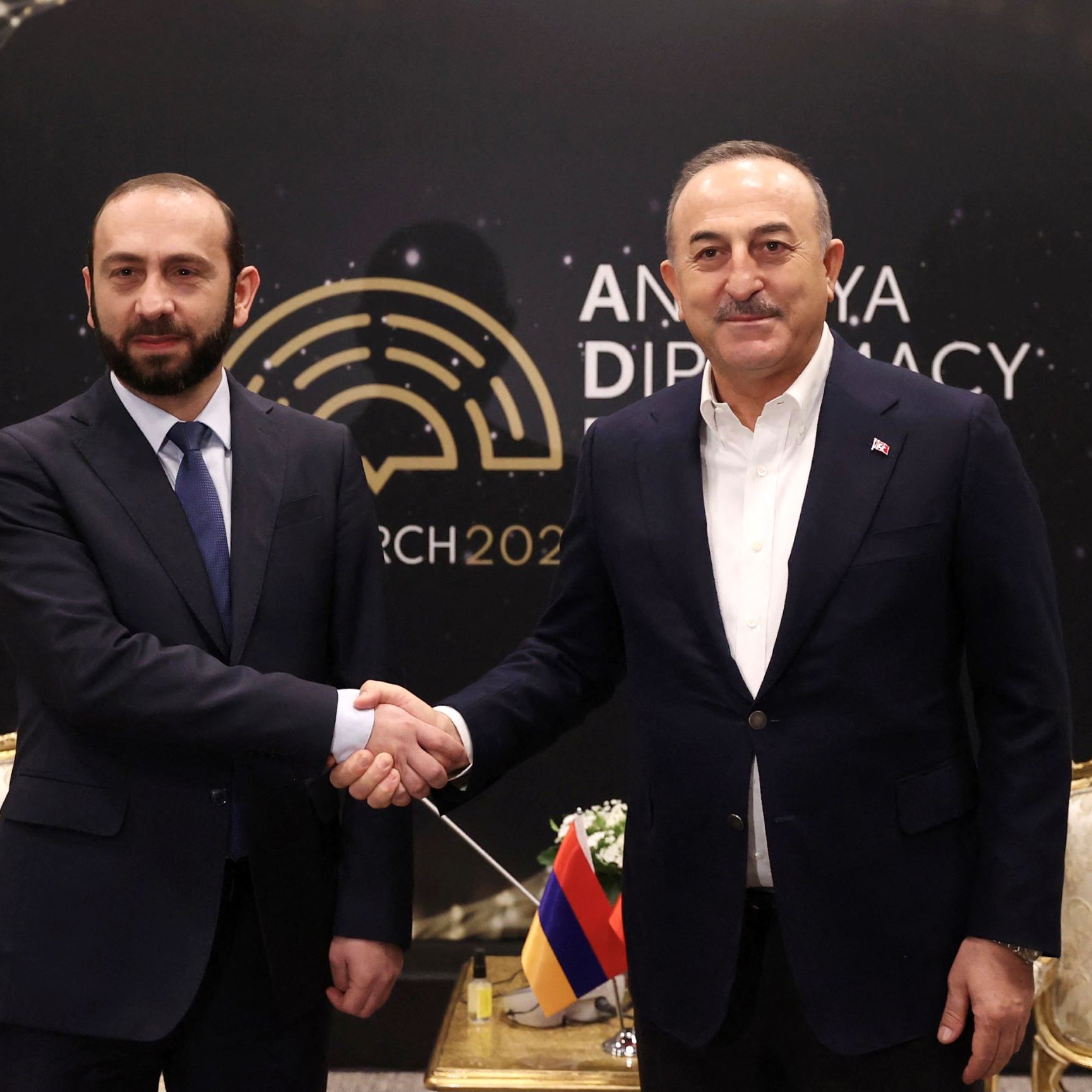 L'image montre deux hommes se serrant la main, probablement en signe de coopération ou de négociation. Ils sont vêtus de costumes formels. L'arrière-plan présente le logo d'un événement lié à la diplomatie, avec la mention "Ankara Diplomacy 2023". L'ambiance semble être celle d'une rencontre officielle.