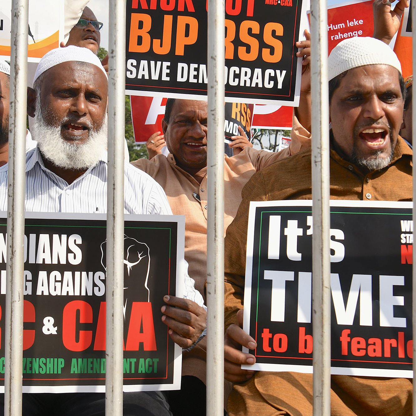 L'image montre un groupe de manifestants qui se tiennent derrière des barreaux. Ils tiennent des pancartes exprimant leur opposition à certaines lois et politiques en Inde, notamment le BJP, le NRC et le CAA. Les manifestants ont des expressions déterminées et sont clairement engagés dans leur cause, appelant à la démocratie et à la résistance. Les pancartes portent des messages en anglais et en d'autres langues, soulignant leur solidarité et leur demande de justice.