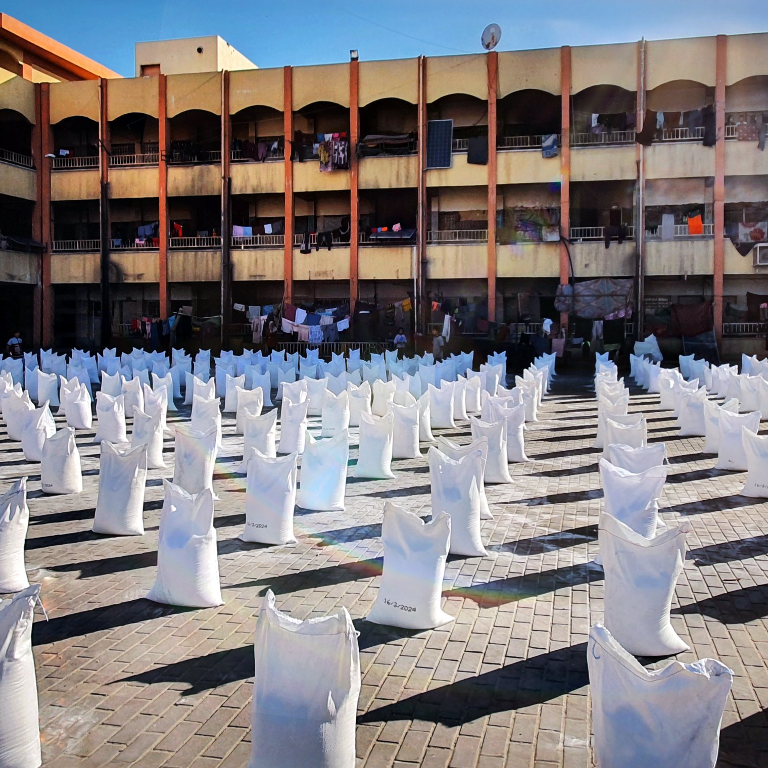 L'immagine mostra un'area all'aperto con molti sacchi bianchi disposti in file ordinate. Sullo sfondo si vede un edificio di più piani, con diversi balconi. Alcuni panni sono appesi ad asciugare tra i balconi. La scena è illuminata dalla luce del sole, creando un'atmosfera vivace e attiva. Il pavimento è costituito da mattonelle in cui si possono notare i segni del tempo. L'insieme suggerisce un contesto urbano con attività legate alla distribuzione di beni.