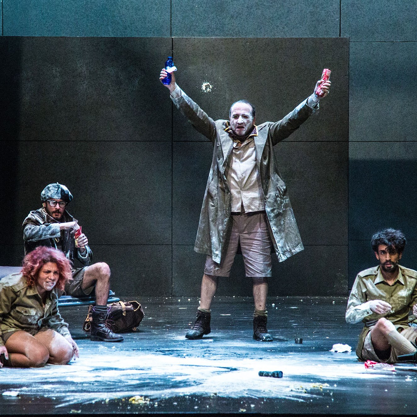 L'image montre une scène de théâtre sur une scène dépouillée. Au centre, un homme en manteau se tient avec les bras levés, tenant une bouteille dans chaque main, ce qui suggère une attitude triomphante ou exubérante. Autour de lui, plusieurs autres personnages sont assis ou accroupis, vêtus de costumes similaires. L'un d'eux semble concentré sur une activité au sol. La scène peut évoquer une ambiance dramatique ou absurde, avec des éléments de désordre comme de la poudre dispersée sur le sol. Les expressions des personnages varient, ajoutant à la dynamique de l'ensemble.