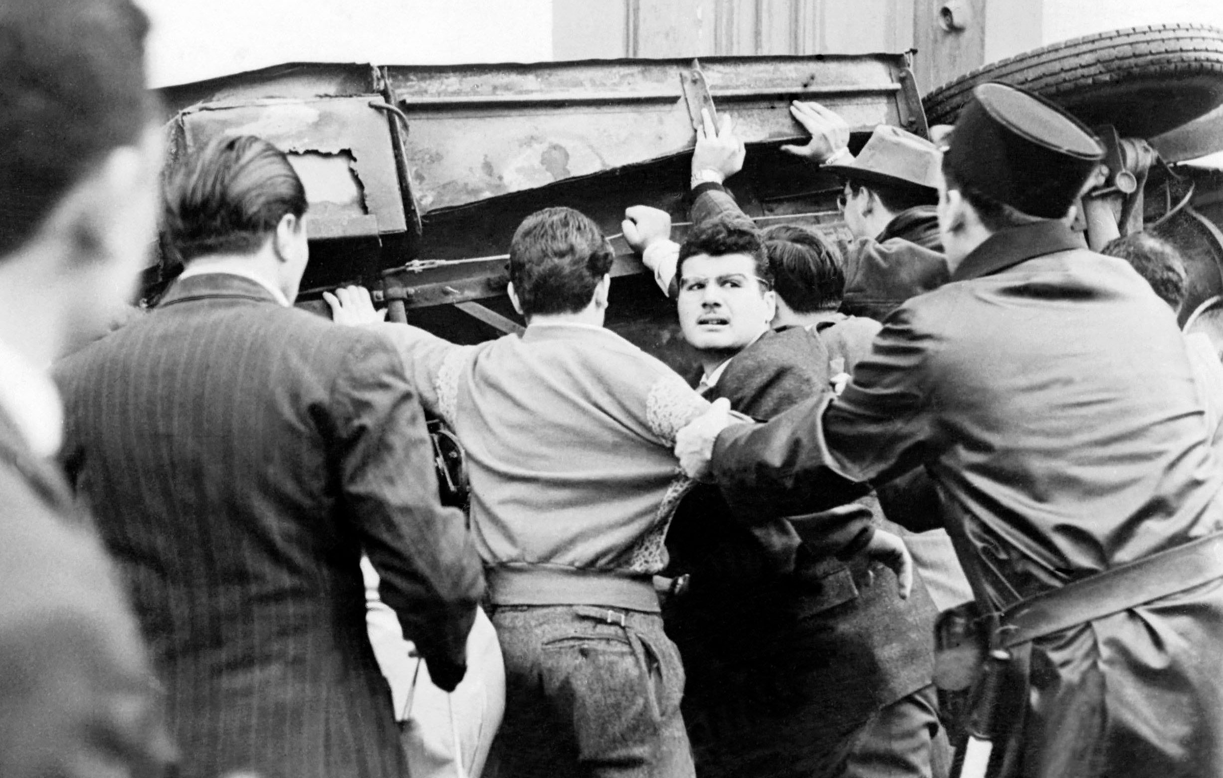 L'image montre une scène tumultueuse où un groupe de personnes semble être impliqué dans une altercation. Au centre, plusieurs hommes s'efforcent de soulever un véhicule renversé, tandis qu'un homme au visage concentré semble être en train de s'opposer à la situation ou d'interagir avec les autres. À droite, un policier en uniforme semble gérer la situation. L'atmosphère est tendue, et le contraste en noir et blanc accentue l'intensité de la scène.