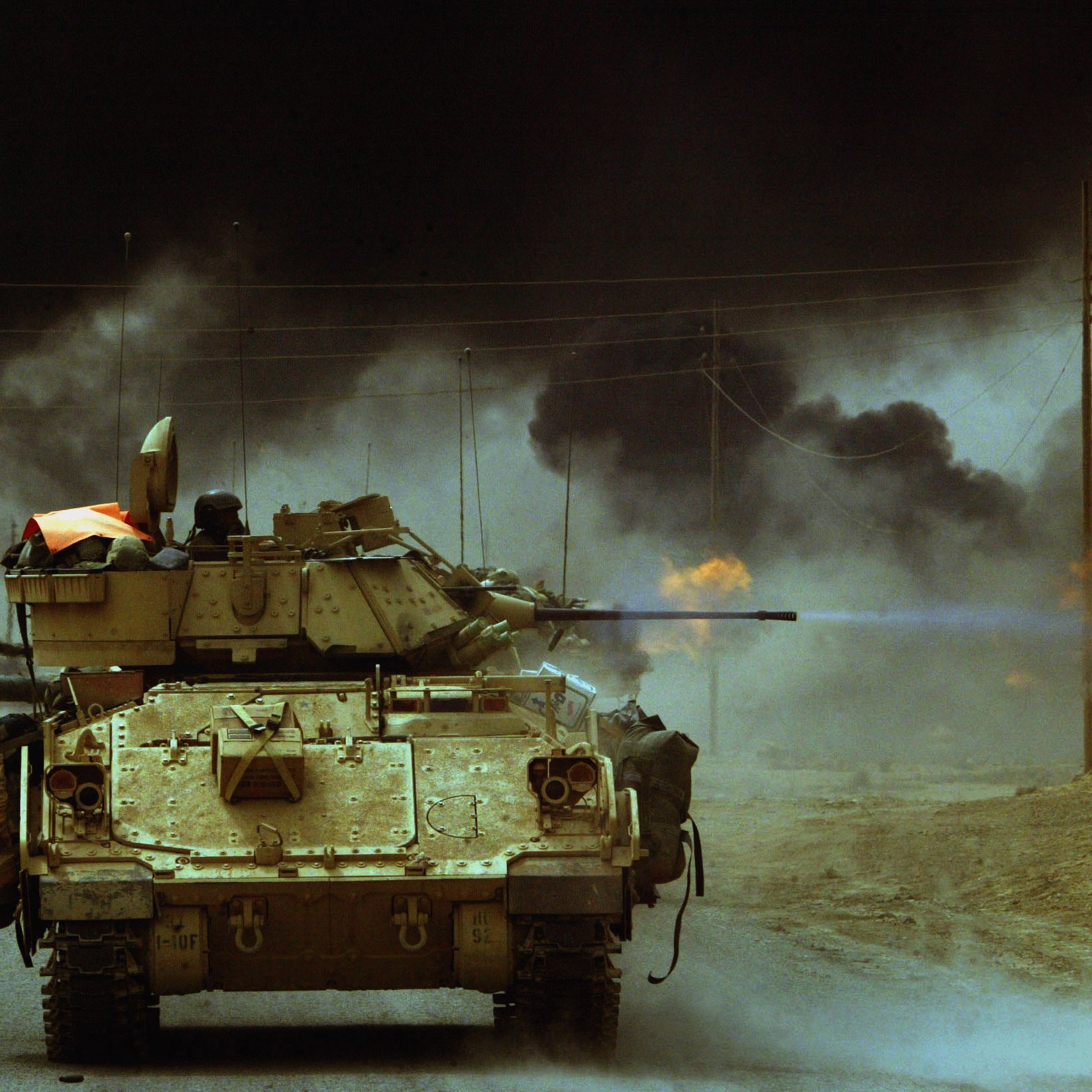 L'image montre un char de guerre sur une route, se dirigeant vers l'avant. En arrière-plan, on aperçoit des nuages de fumée noire qui s'échappent de flammes, suggérant un environnement de conflit ou d'escalade militaire. La scène est sombre et dramatique, évoquant une ambiance de tension et de danger. Le char est équipé d'un canon, et l'ensemble de l'image dépeint une situation de guerre.