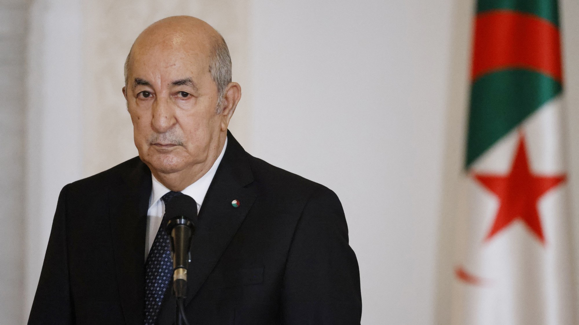 L'image montre un homme en costume sombre, regardant vers l'avant avec une expression sérieuse. Il se tient devant un microphone, et en arrière-plan, on peut voir un drapeau. Le drapeau présente des couleurs vertes et rouges avec une étoile et un croissant. L'ambiance de l'image semble solennelle, suggérant un contexte formel ou officiel.