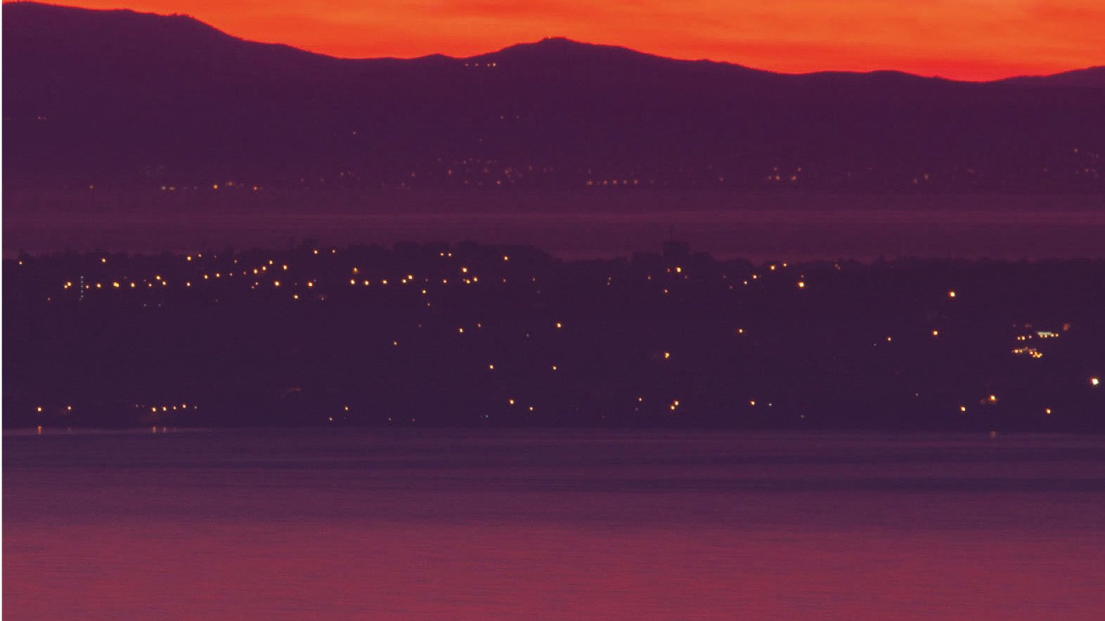 L'image présente un coucher de soleil spectaculaire, où le ciel est teinté de rouge et d'orange lumineux. En arrière-plan, on aperçoit des collines sombres, tandis qu'à l'avant-plan, un cargo navigue sur l'eau calme. Les lumières d'une ville ou d'un port scintillent le long de la côte, ajoutant une touche de vie à cette scène paisible.