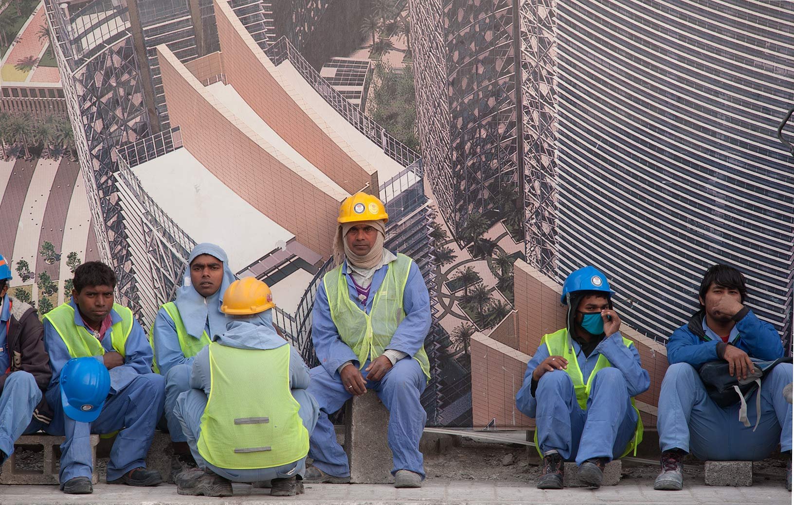 En la imagen se pueden ver a varios trabajadores de la construcción sentados sobre una superficie. Están vistiendo uniformes azules y chalecos reflectantes. Algunos llevan casco de diferentes colores, como amarillo y azul. Al fondo, hay un gran cartel que muestra una imagen de un edificio moderno, sugiriendo que están trabajando en un proyecto de construcción. Los trabajadores parecen estar en un descanso, y el ambiente refleja una jornada laboral intensa.