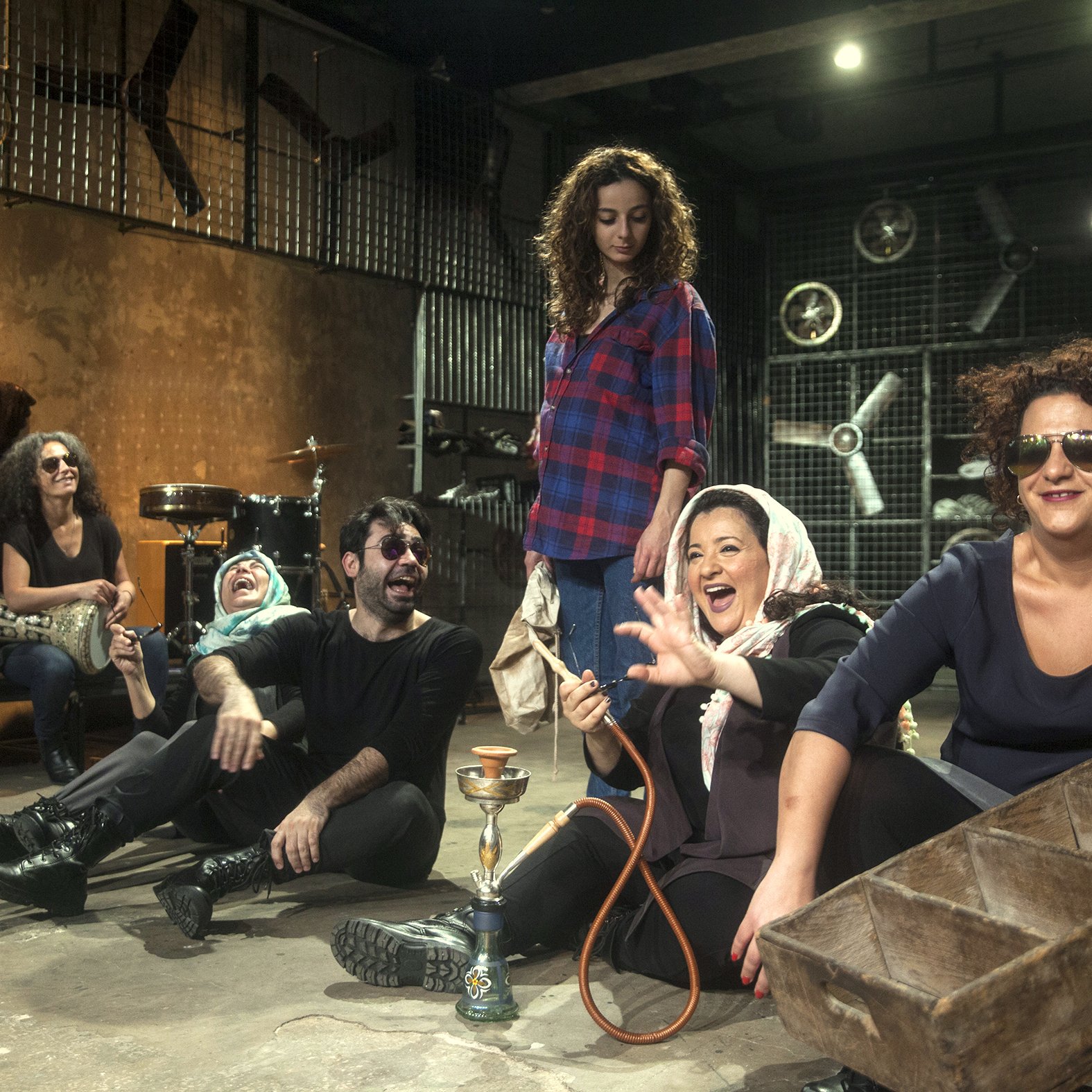 L'image présente un groupe de personnes assises sur le sol d'un espace décoratif et artistique. On y voit des femmes et des hommes, certains souriant et riant, dans une ambiance décontractée et joyeuse. Les femmes portent des vêtements variés, y compris un hijab, tandis que certains d'entre eux portent des lunettes de soleil. À l'arrière-plan, il y a des instruments de musique et des éléments de décor industriels, ce qui ajoute une touche créative à la scène. Au centre, une femme tient une chicha (narguilé) et semble engagée dans la conversation. L'ensemble de la composition donne une impression de camaraderie et de convivialité.