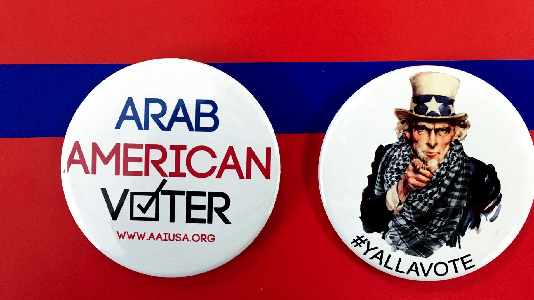 الصورة تحتوي على زرين دائرين. الزر الأول مكتوب عليه "ARAB AMERICAN VOTER" مع علامة التحقق، مما يدل على التصويت للمواطنين العرب الأمريكيين. الزر الثاني يظهر صورة لشخص يرتدي قبعة تحمل نجمة، وهو رمز معروف في الثقافة الأمريكية، مع عبارة "#YALLAVOTE" التي تدعو الناس للتصويت. الخلفية حمراء مع شريط أزرق، مما يضيف طابعاً احتفالياً للموضوع.