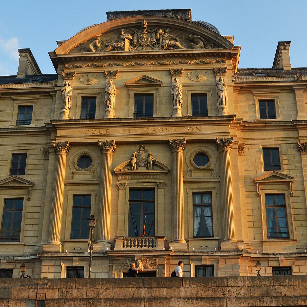L'image montre un bâtiment imposant, probablement un palais ou un tribunal, éclairé par la lumière dorée du soleil couchant. La façade est ornée de détails architecturaux élaborés, avec des colonnes et des sculptures qui ajoutent à son majestueux style classique. Le nom "COUR DE CASSATION" est visible, indiquant qu'il s'agit d'un lieu judiciaire important. Le cadre montre un ciel clair, accentuant la beauté de l'architecture historique.