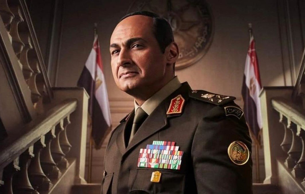 يظهر في الصورة رجل يرتدي زيًا عسكريًا يحمل العديد من الأوسمة والرتب. يبدو أنه يتخذ وضعًا رسميًا وجادًا، مع خلفية تتضمن علمين. تعكس تعابير وجهه الثقة والسلطة، كما أن التصميم العام للصورة يوحي بأجواء عسكرية أو حكومية.