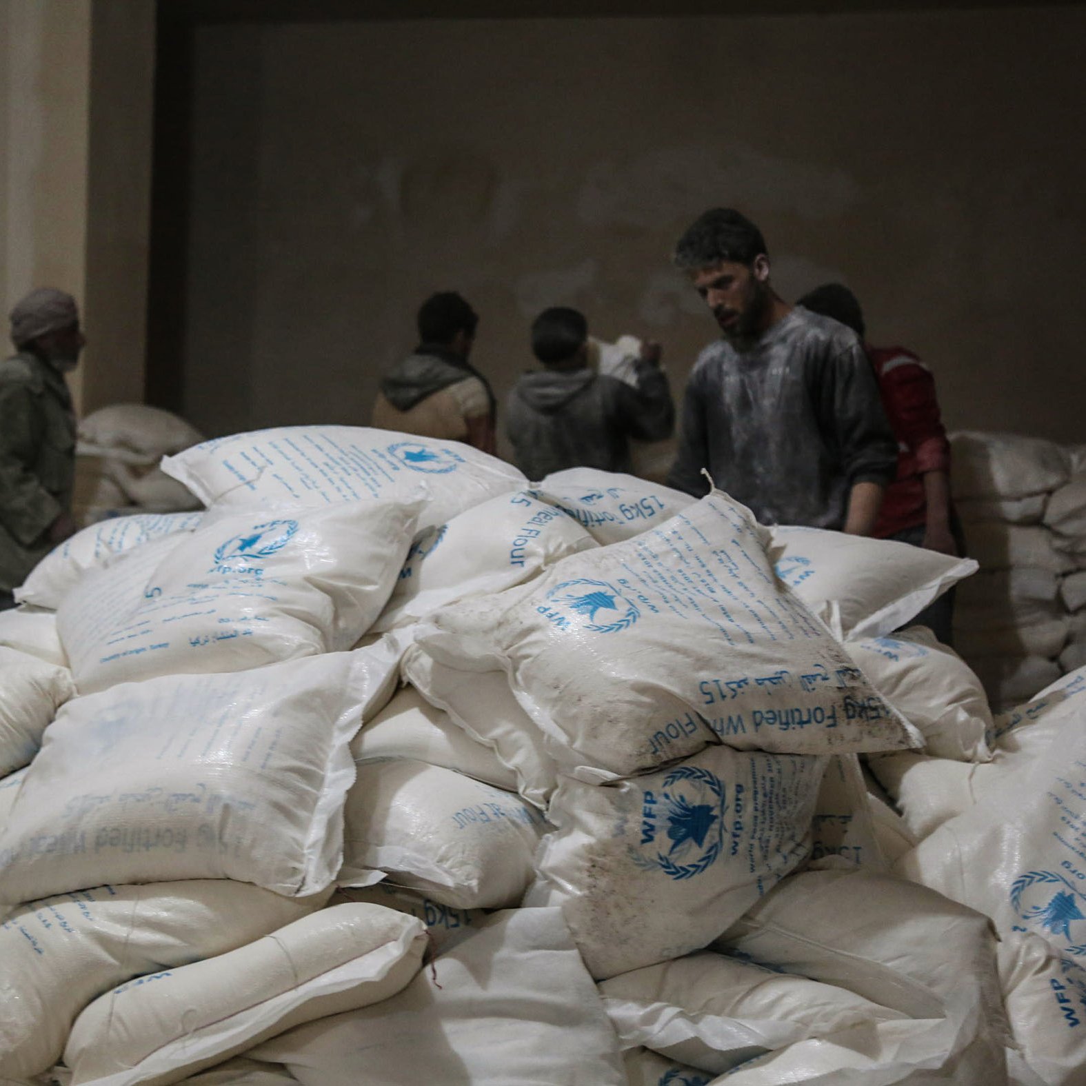 تظهر في الصورة مجموعة من الأشخاص داخل مكان يبدو أنه مخزن للمواد الإغاثية. في المقدمة، هناك كومة كبيرة من الأكياس البيضاء التي تحمل علامات معينة، ويبدو أن الأكياس تحتوي على مواد غذائية أو إغاثية. في الخلفية، يمكن رؤية بعض الأشخاص الذين يتحدثون أو يعملون. الإضاءة في المكان خافتة، مما يعطي شعورًا بالجدية والاهتمام بالعمل الذي يقومون به.