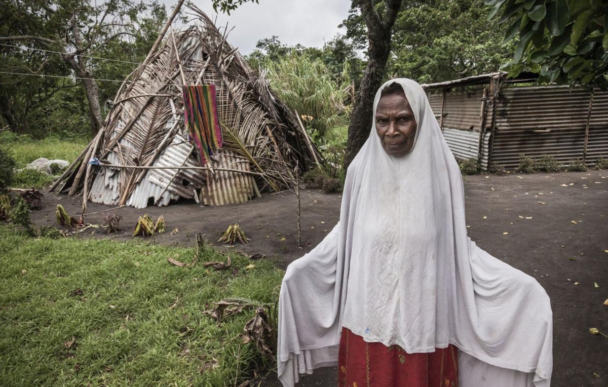 Sur cette image, on voit une femme debout au premier plan, portant un vêtement traditionnel avec un voile qui couvre sa tête. Son expression semble sérieuse ou réfléchie. À l'arrière-plan, on aperçoit une structure habitable, partiellement en bois et en matériaux naturels, qui semble être endommagée. La scène est entourée de verdure, avec des arbres et de l'herbe. L'ambiance générale évoque un contexte rural ou tribal, suggérant une connexion forte avec la nature et un mode de vie simple.