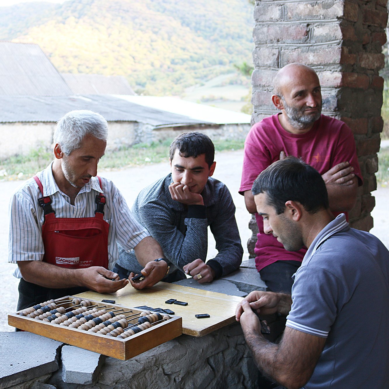 تصویر گروهی از مردان را نشان می‌دهد که در یک محیط روستایی گرد هم آمده‌اند. در مرکز تصویر، مردی با موهای سفید و لباس‌های کارگاهی به نظر می‌رسد که مشغول توضیح یا نشان دادن چیزی به دیگران است. مردان دیگر به دقت به او گوش می‌دهند و در حال بررسی یک صفحه بازی هستند که تعدادی مهره در آن قرار دارد. پس‌زمینه تصویر شامل تپه‌های سرسبز و یک ساختمان قدیمی است. جو عمومی تصویر دوستانه و غیررسمی به نظر می‌رسد.