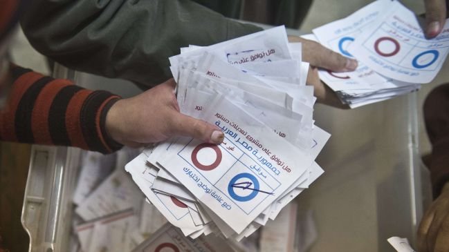 L'image montre des mains tenant des bulletins de vote. On peut voir des bulletins blancs portant des cercles rouges et bleus, ainsi que des inscriptions en arabe. Il semble s'agir d'une scène de vote, probablement lors d'une élection. Les bulletins sont rassemblés, suggérant un processus électoral en cours.