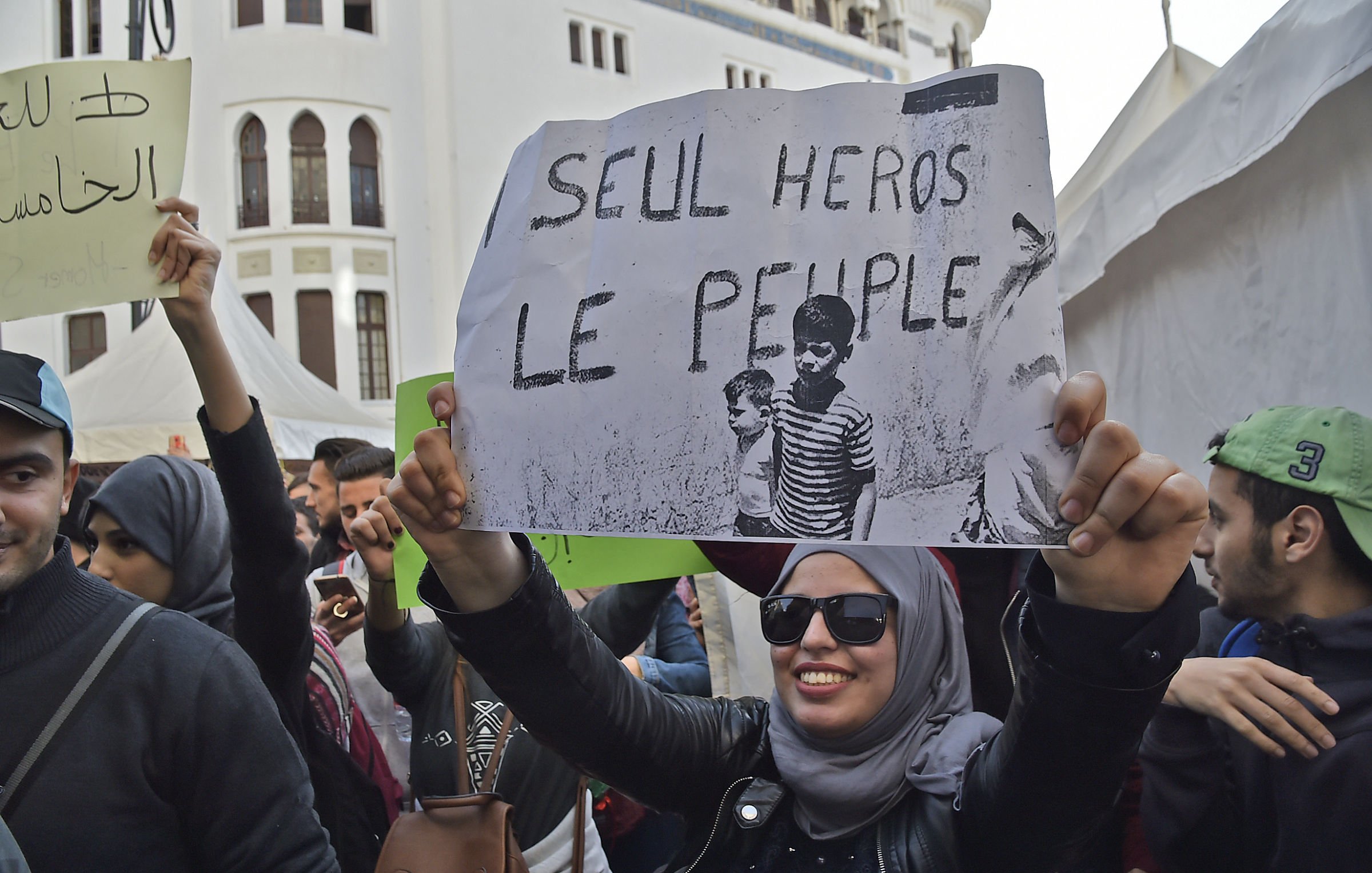 در این تصویر، تعدادی از مردم در یک تجمع حضور دارند. فردی جلوی دوربین ایستاده و یک پلاکارد بزرگ را در دست دارد که روی آن نوشته «SEUL HÉROS LE PEUPLE؟» به معنای «تنها قهرمان مردم؟» دیده می‌شود. فردی که پلاکارد را در دست دارد، عینک دودی به چشم دارد و روسری به سر دارد. اطراف او جمعیت متنوعی از افراد قابل مشاهده است که احتمالاً در حال مشارکت در یک رویداد اجتماعی یا اعتراض هستند. پس‌زمینه تصویر شامل سازه‌های معماری و چادرهایی است که به نظر می‌رسد برای این تجمع برپا شده‌اند.