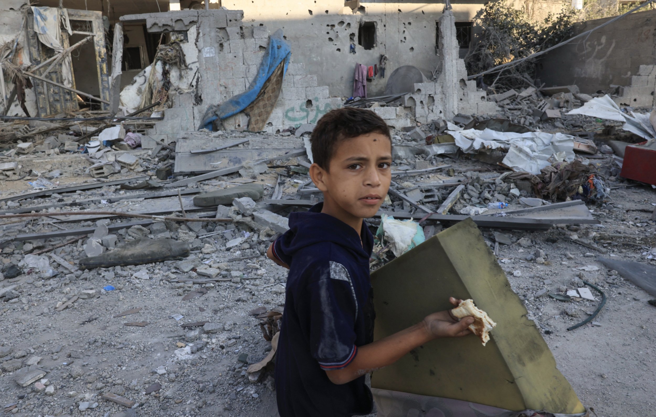 L'image montre un jeune garçon au visage marqué par l'environnement chaotique autour de lui. Il porte des vêtements simples et tient un morceau qu'il semble avoir trouvé dans les débris. Le fond est constitué de bâtiments détruits, avec des murs endommagés et des débris éparpillés, ce qui évoque une situation de conflit ou de catastrophe. La lumière du jour éclaire la scène, mettant en évidence la gravité de la situation dans laquelle il se trouve.