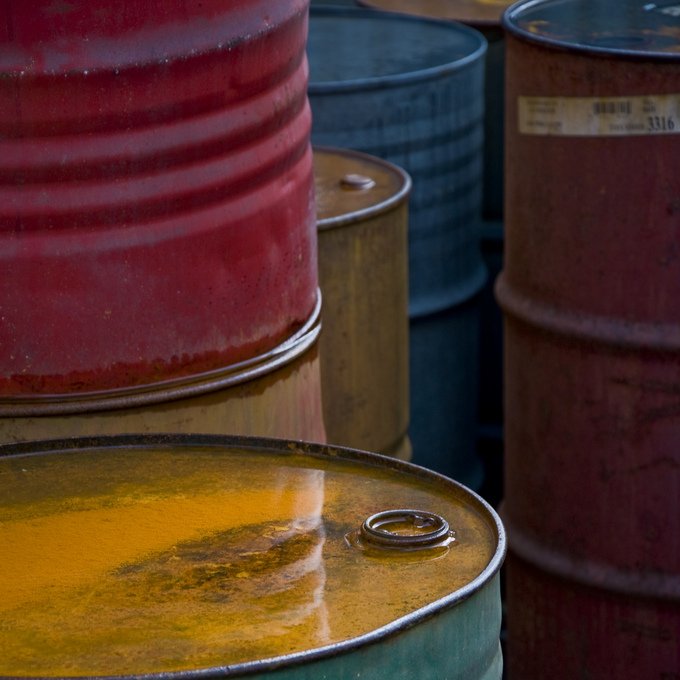 L'image montre plusieurs fûts en métal empilés les uns sur les autres. Chaque fût a une couleur différente : rouge, vert, noir et jaune. La surface des fûts semble briller, probablement à cause d'un liquide qui s'y trouve, reflétant la lumière. L'ensemble de la composition donne une impression de dépôt industriel, avec une texture rustique et des nuances de couleurs intéressantes.