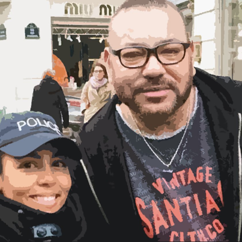 L'image montre deux personnes posant ensemble à l'extérieur. L'une porte une casquette avec le mot "POLICE" et a un sourire, tandis que l'autre, à la barbe bien taillée et portant des lunettes, a une expression amicale. L'arrière-plan montre une zone commerçante animée avec des gens et des boutiques.