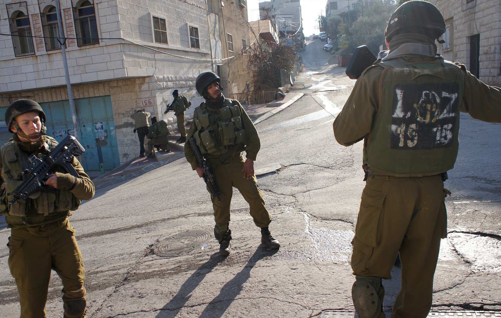 L'image montre des soldats armés en tenue militaire dans une rue apparemment urbaine. Deux d'entre eux sont en position de vigilance, tandis qu'un autre semble communiquer ou surveiller. L'environnement semble être un quartier situé dans une ville, avec des bâtiments en arrière-plan. L'atmosphère pourrait suggérer une situation de tension ou de surveillance.