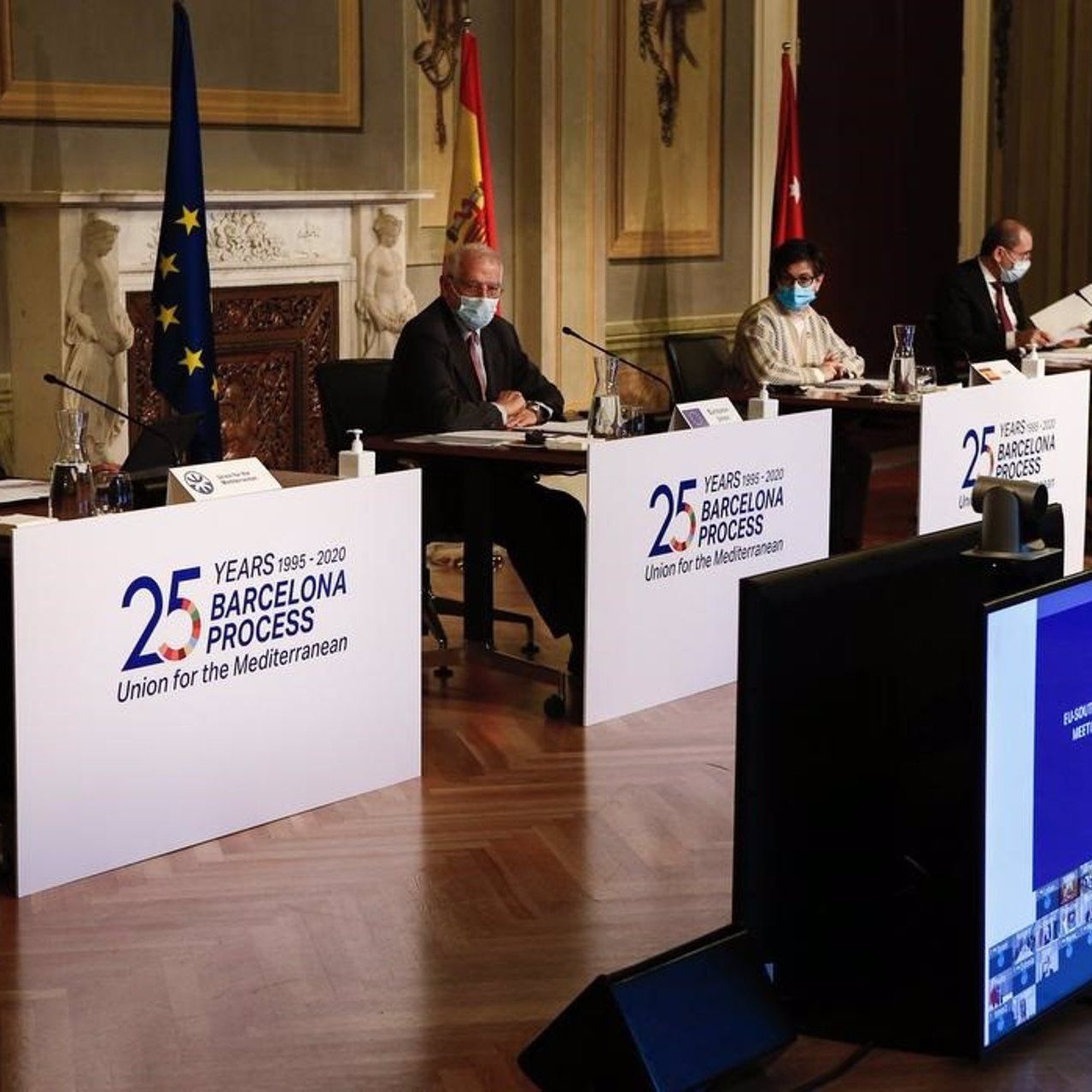 L'image montre une scène de conférence avec plusieurs intervenants assis derrière des panneaux portant le logo du processus de Barcelone, qui célèbre ses 25 ans. Les participants portent des masques, indiquant qu'ils suivent des mesures de santé, probablement en raison de la pandémie. À l'arrière-plan, on peut voir des drapeaux représentant l'Union européenne et l'Espagne, ainsi qu'un écran affichant des images, probablement liées à l'événement. L'environnement semble formel, suggérant une réunion diplomatique ou un sommet.