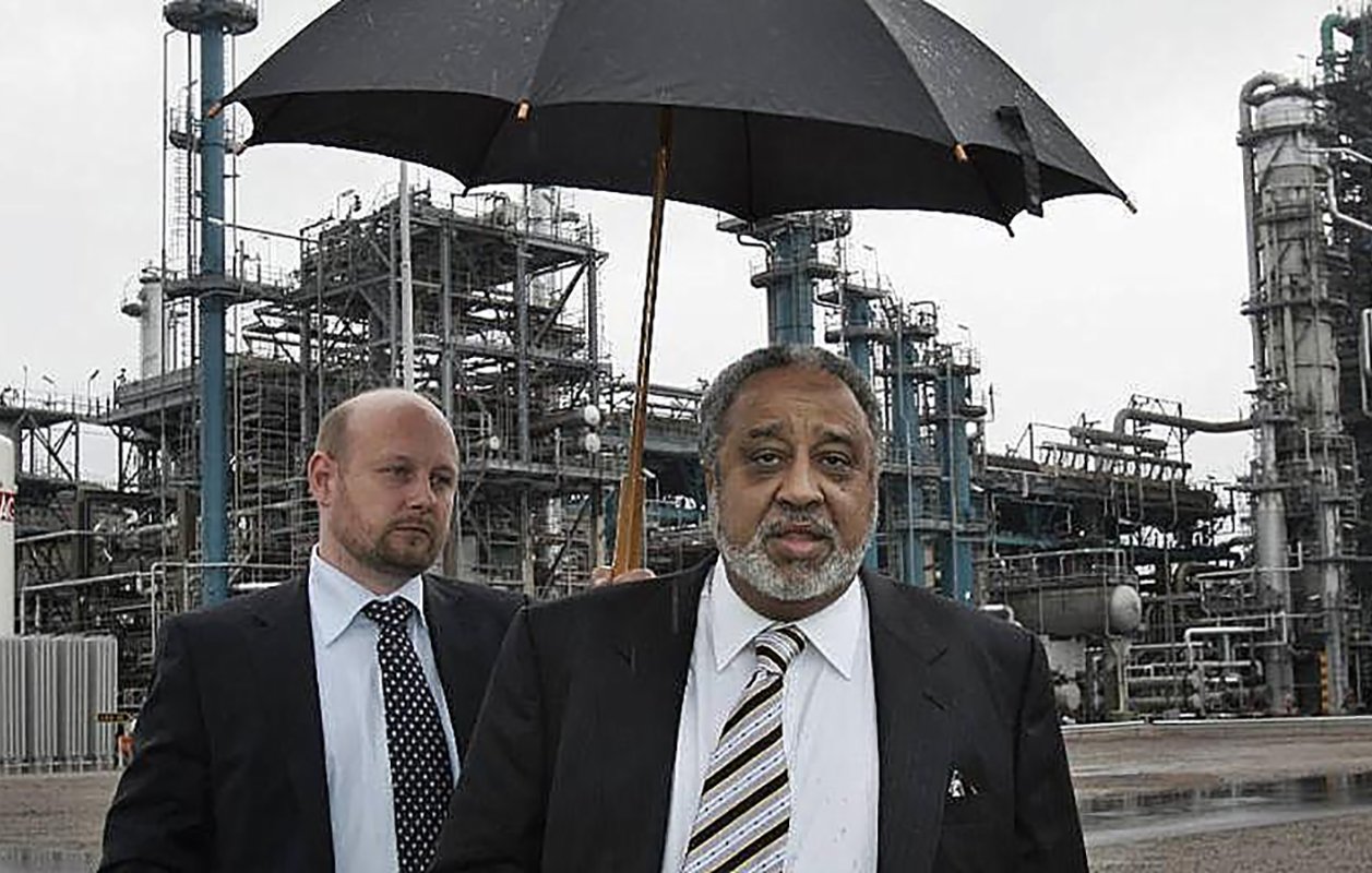 الصورة تظهر رجلين في موقع صناعي. الرجل في المقدمة يحمل مظلة سوداء، ويبدو أنه يتحدث أو يتفاعل مع شخص آخر. الرجل الثاني يقف خلفه بنظرة جدية، ويرتدي بدلة رسمية. في الخلفية، توجد منشآت صناعية تظهر أنابيب ومعدات، مما يشير إلى أن المكان هو مصنع أو منشأة صناعية. السماء غائمة، مما يدل على أنه قد يكون هناك مطر.