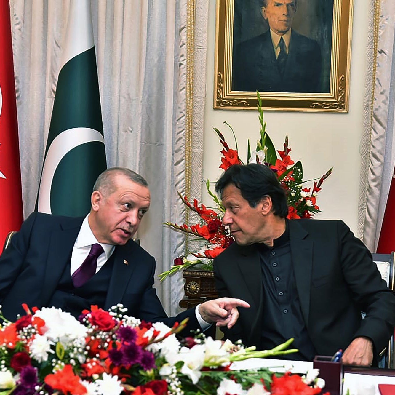 تظهر الصورة اجتماعًا بين شخصيتين بارزتين. يجلس الرجلان على طاولة مزينة بورود ملونة. خلفهما علم تركيا وعلم باكستان، بالإضافة إلى صورة لشخصية تاريخية معلقة على الجدار. يبدو أن الرجلين يتبادلان الحديث بشكل ودي، حيث يشير أحدهما بإصبعه كما لو كان يشرح شيئًا مهمًا. الإضاءة في الغرفة دافئة وتضفي جوًا من الألفة على الاجتماع.