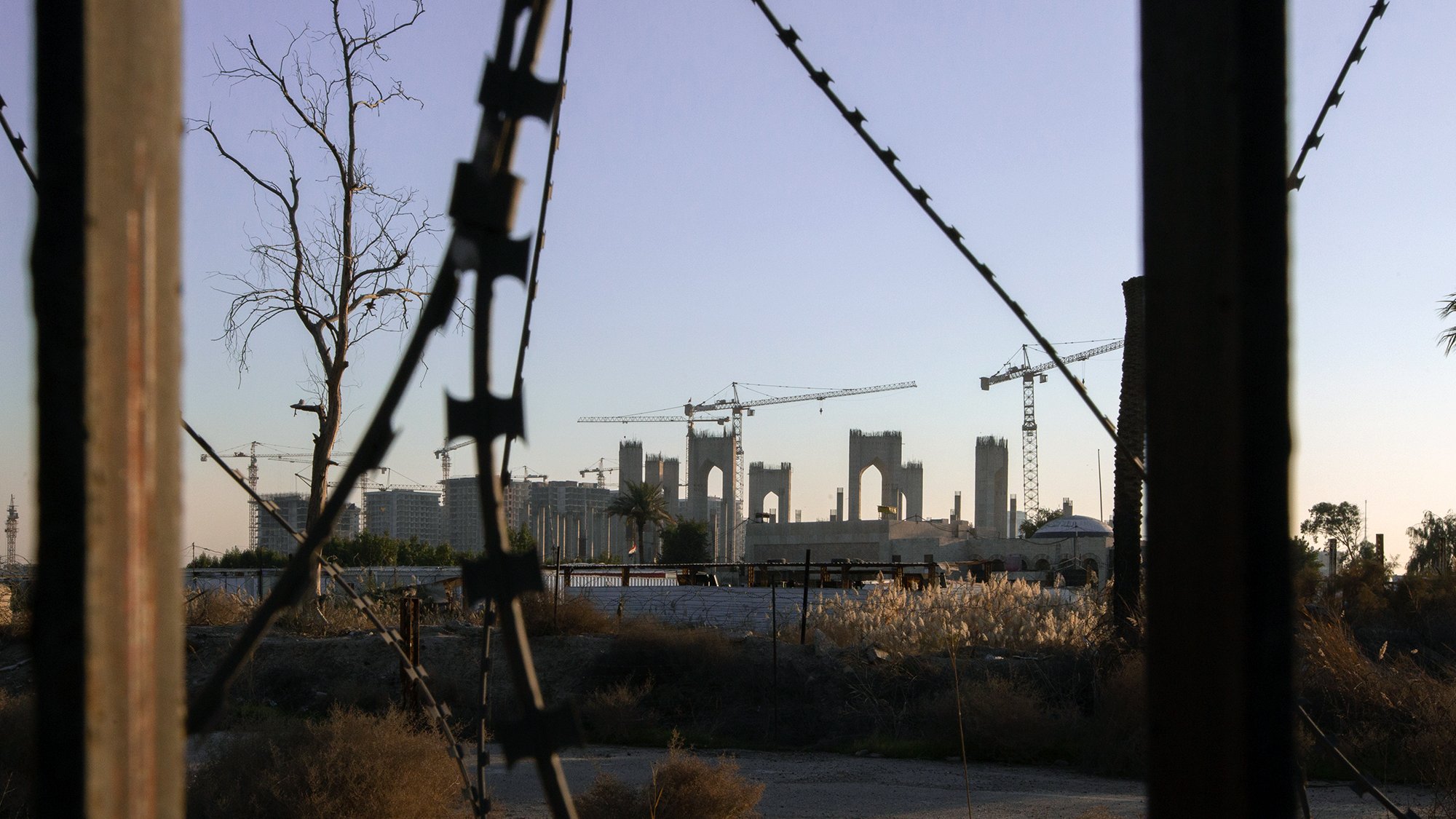 L'image présente un paysage urbain en transformation, avec des structures inachevées à l'arrière-plan, entourées de grues de construction. En premier plan, on peut voir des barbelés et des éléments de végétation desséchée, suggérant un environnement abandonné ou en déclin. La lumière douce du crépuscule ajoute une ambiance mélancolique à la scène, mettant en contraste l'espoir de la construction avec la désolation du terrain environnant.