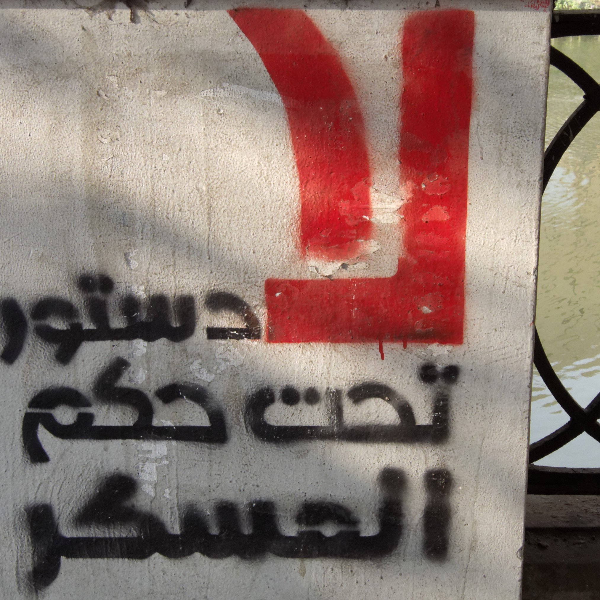 L'image montre un panneau sur un mur en béton, avec un fond gris. Sur la partie supérieure, on peut voir un symbole ou une lettre en rouge. En dessous, des mots sont écrits en arabe, et le texte semble faire référence à un sujet politique, évoquant un appel ou une déclaration. Le tout est en contraste avec l'environnement aquatique en arrière-plan.