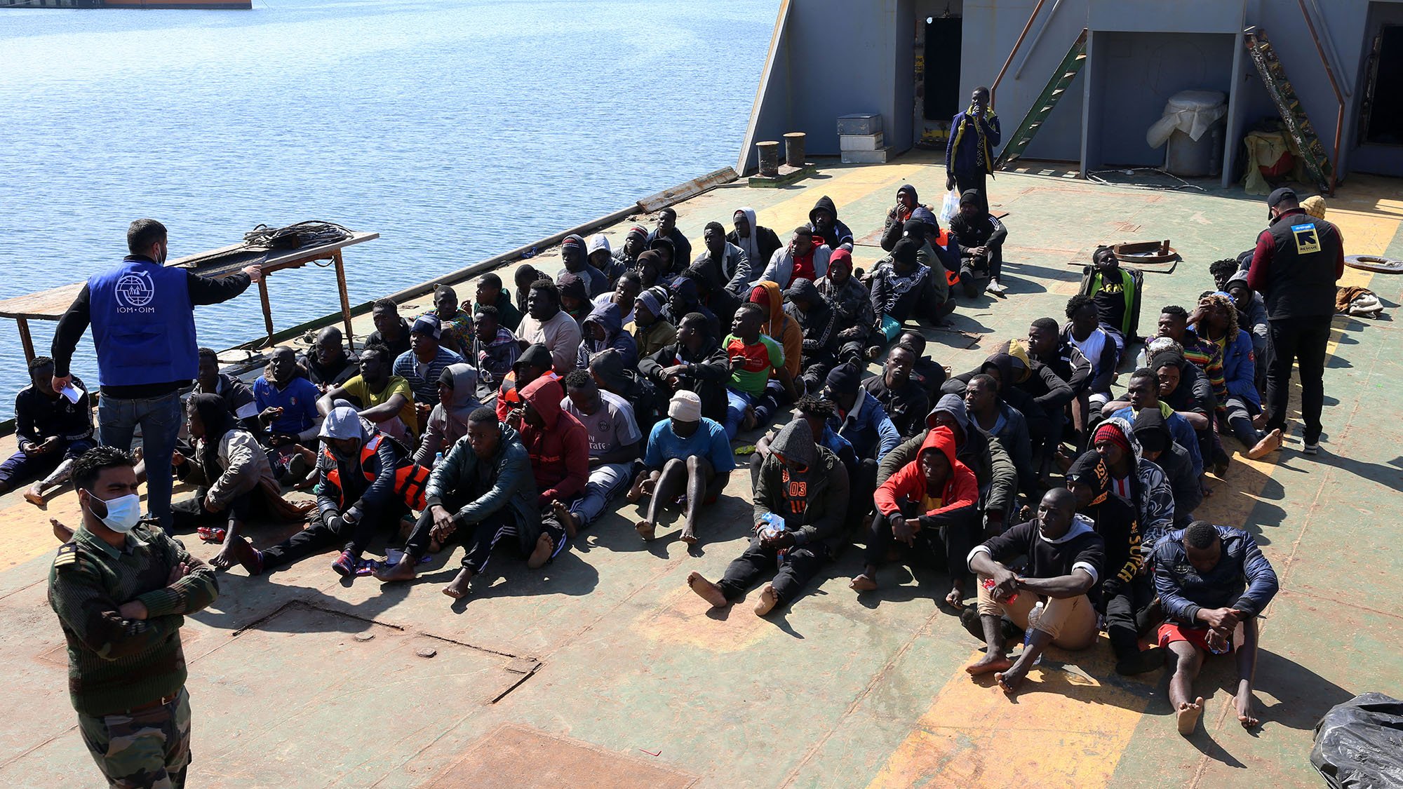 در این تصویر، گروهی از افراد در یک کشتی نشسته‌اند. آنها در حالتی متراکم و منظم روی عرشه نشسته‌اند و بیشتر آنها لباس‌های گرم به تن دارند. برخی از آنها هم کلاه و ماسک به صورت دارند. در سمت چپ تصویر، فردی با یونیفورم نظامی و شخصی دیگر با جلیقه آبی در حال نظارت بر این گروه هستند. در پس‌زمینه، آب دریا و بخشی از ساحل قابل مشاهده است. جو کلی تصویر به نظر می‌رسد که مربوط به یک عملیات نجات یا مهاجرت باشد.