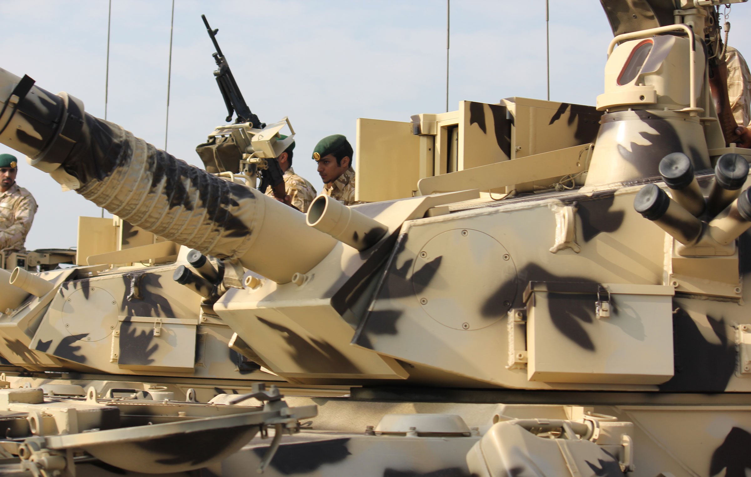 L'image montre un char militaire avec un camouflage dans des tons de beige et de marron. On peut voir un soldat à proximité, portant un uniforme militaire et un casque. Le char est équipé d'un canon en position, et il semble être prêt pour une opération. L'environnement semble être en plein air, peut-être lors d'exercices ou d'une parade militaire, avec un ciel clair en arrière-plan.