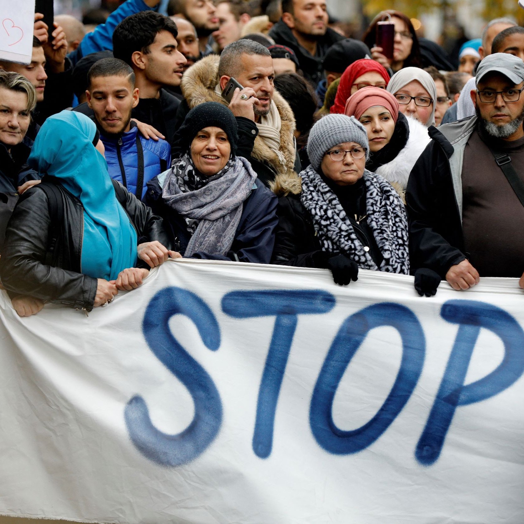 الصورة تظهر مجموعة من الأشخاص الذين يشاركون في مظاهرة. يحتفظ المتظاهرون بلافتة مكتوب عليها كلمة "STOP" بشكل بارز. يتواجد في الصفوف مجموعة متنوعة من المشاركين، حيث يرتدي بعضهم غطاء رأس. يبدو أنهم يعبرون عن موقف معين أو مطلب اجتماعي. الأجواء تشير إلى تواجد حماس وحيوية في العرض، مع انطباع قوي من الوحدة بين الناس.
