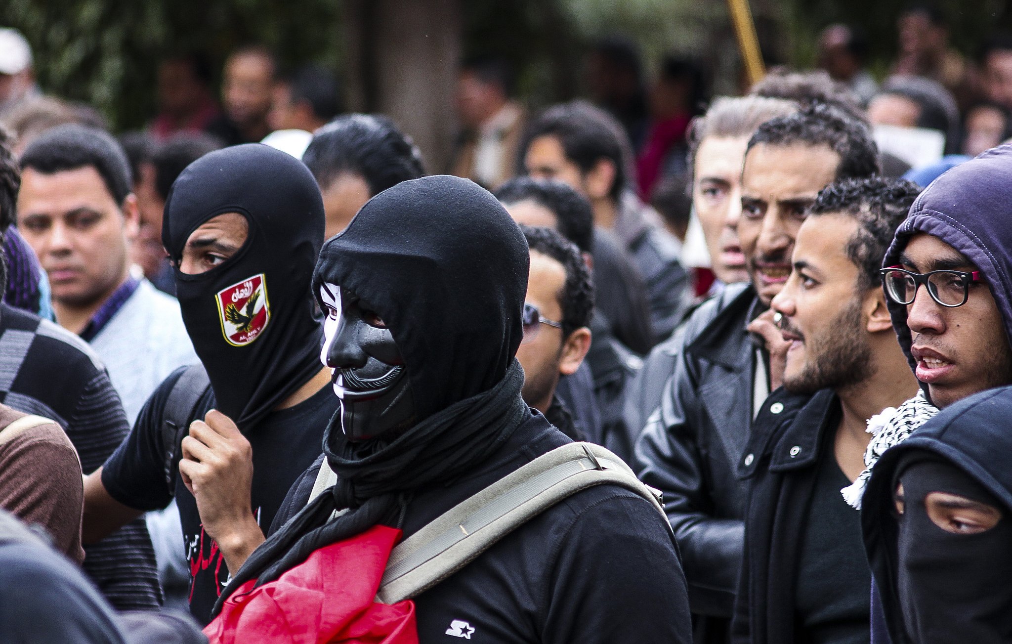 L'image montre un groupe de personnes rassemblées, probablement lors d'une manifestation. Certains portent des masques noirs, tandis que d'autres affichent des expressions déterminées. On peut observer un mélange de vêtements allant des vestes en cuir à des écharpes. L'ambiance semble être à la fois sérieuse et engagée, avec des signes de solidarité entre les participants.